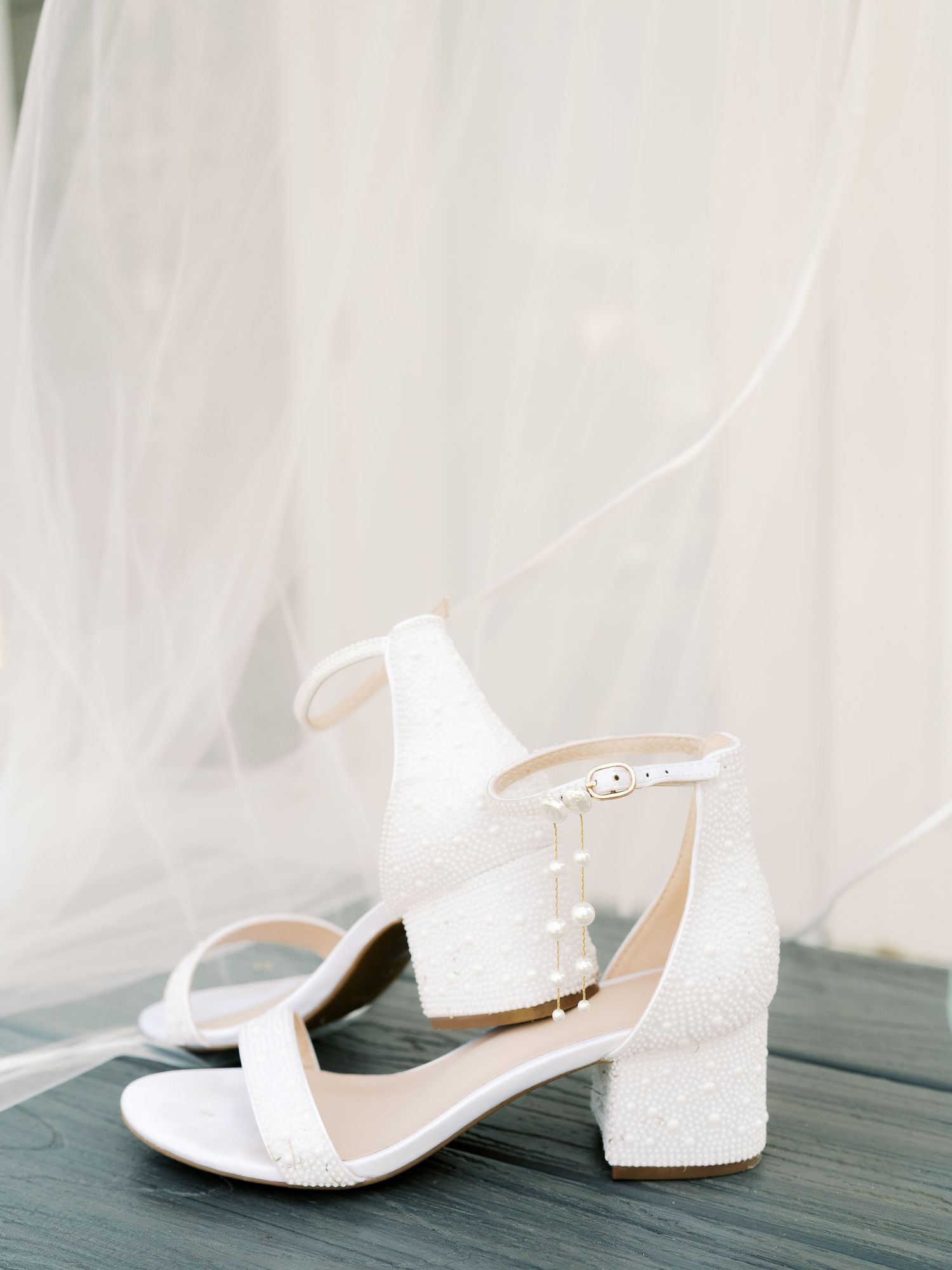 Elegant white bridal shoes with delicate beading and ankle straps displayed on a dark wooden surface.