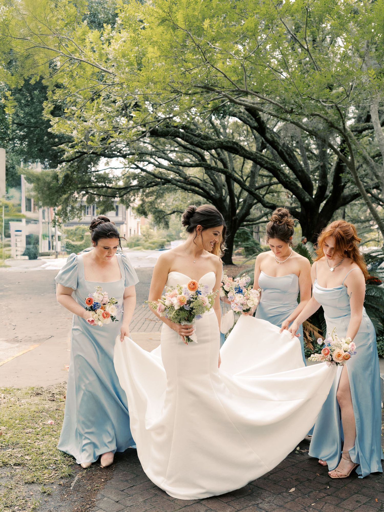 Bridesmaids in light blue dresses help arrange the bride's flowing white dress outdoors.
