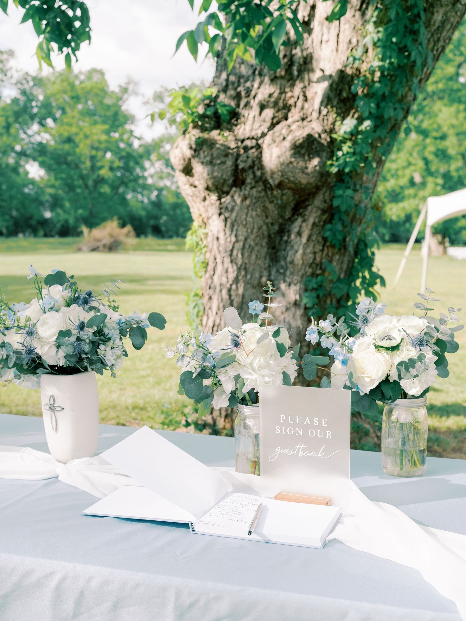 Elegant wedding guest book table with white floral arrangements and greenery under a large tree in an outdoor setting.