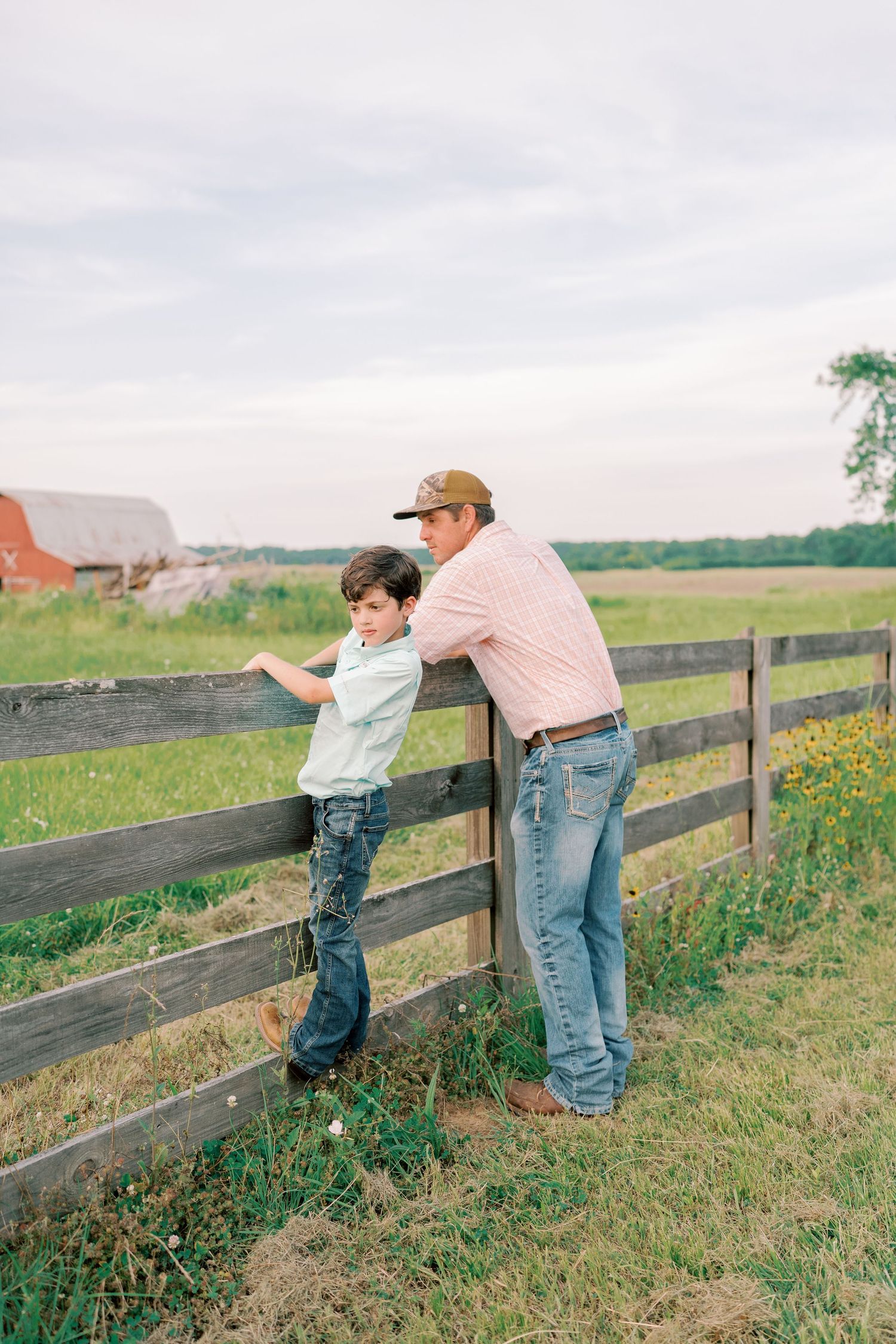 Parent and child exploring along a wooden fence in a grassy countryside setting.