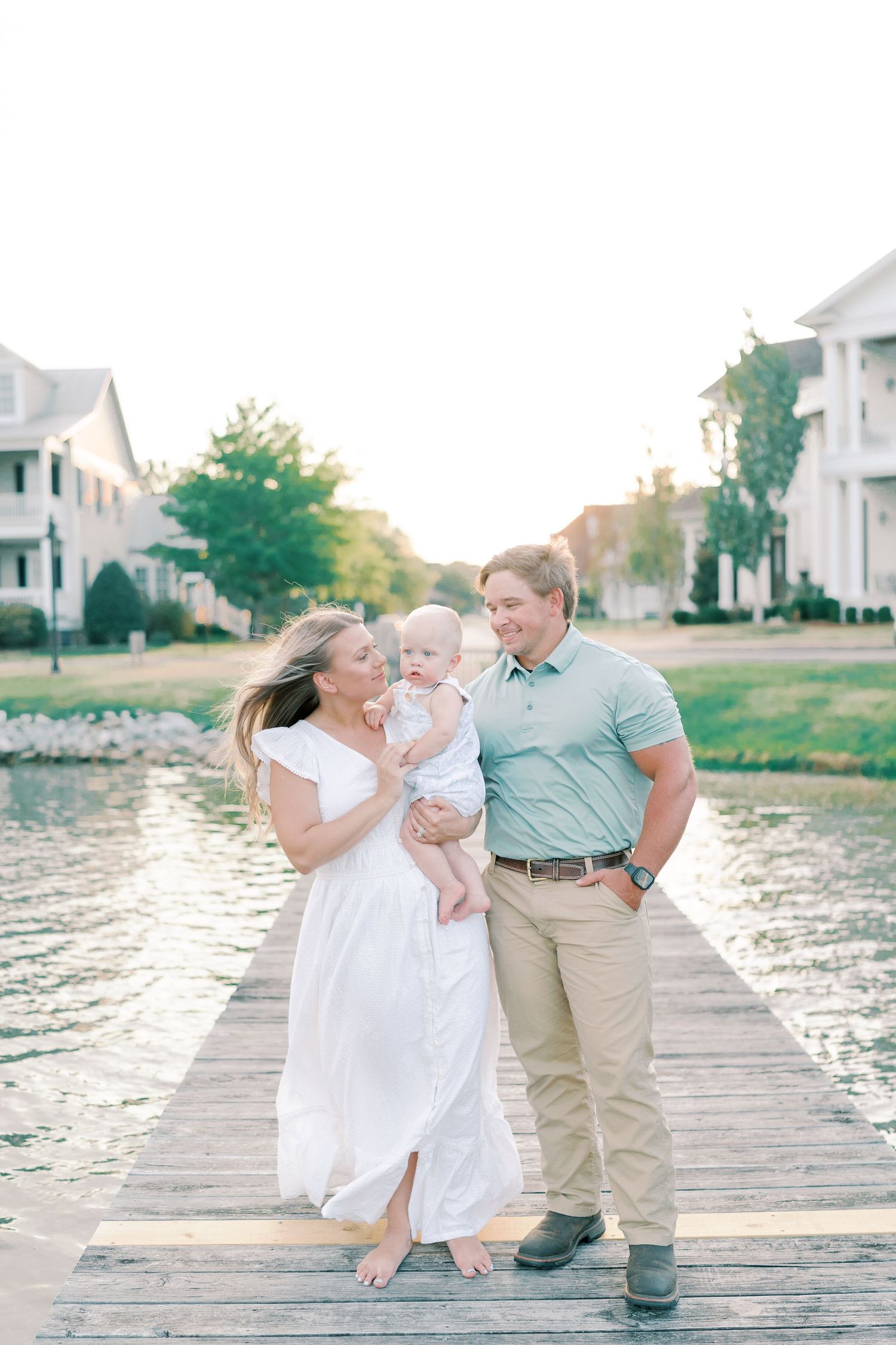 Family dressed in white and khaki sharing loving moments on a lakeside dock at sunset.