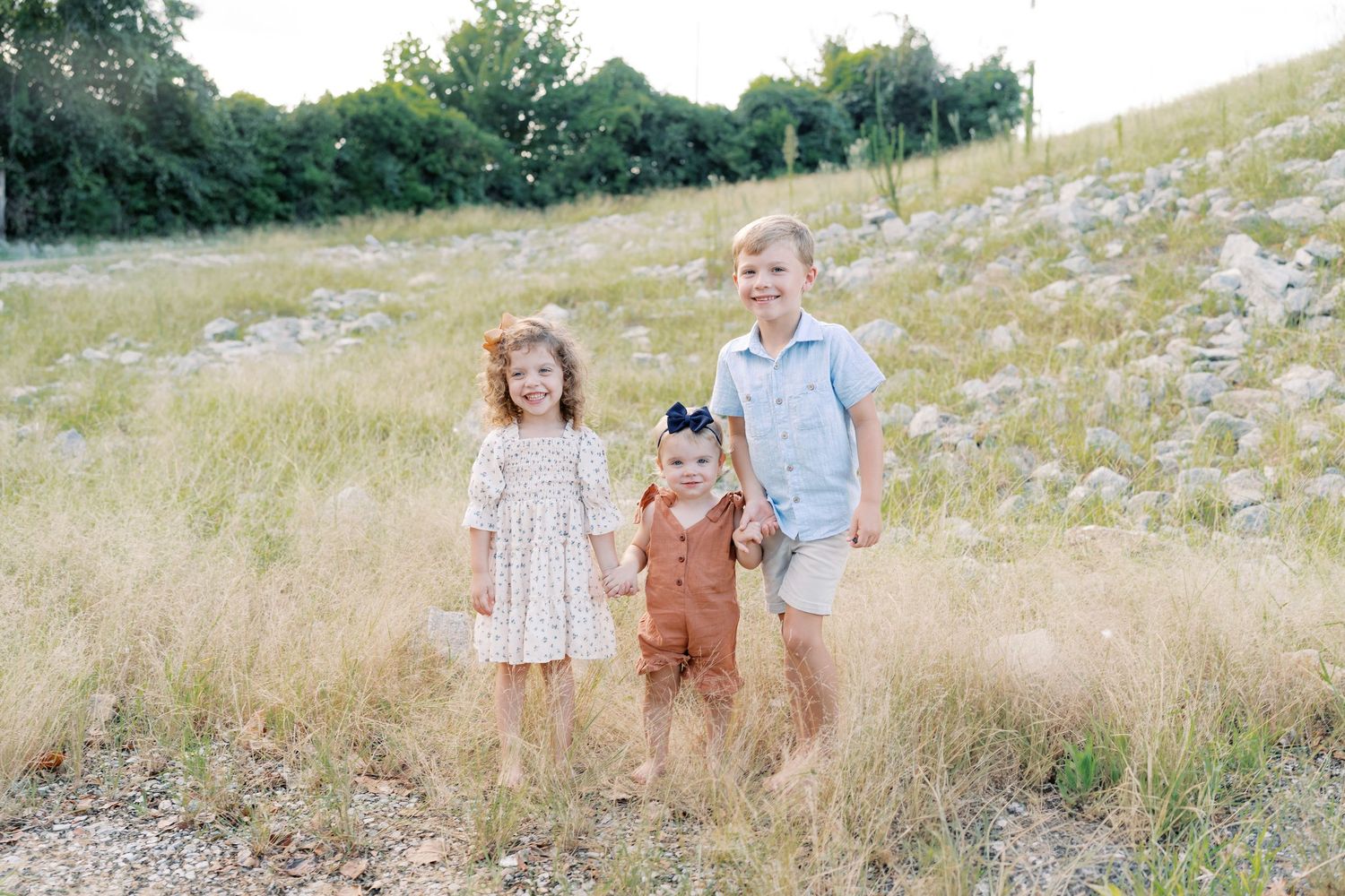 Three siblings standing together in a natural meadow setting with rocks and tall grass.