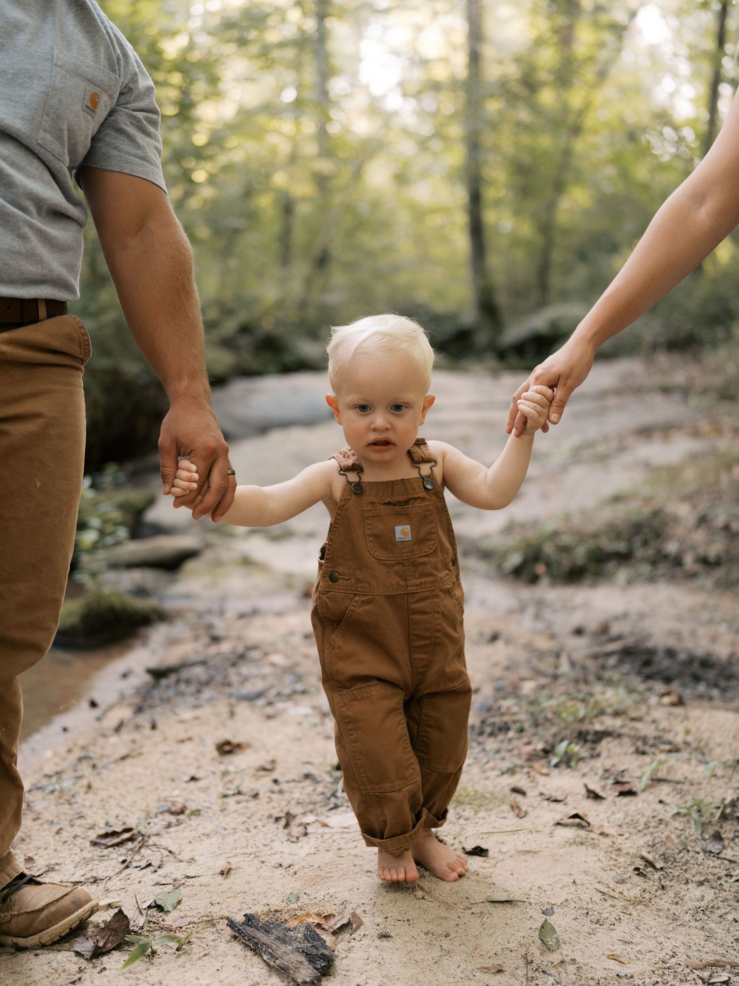 Parents help their baby learn to walk on a forest trail.