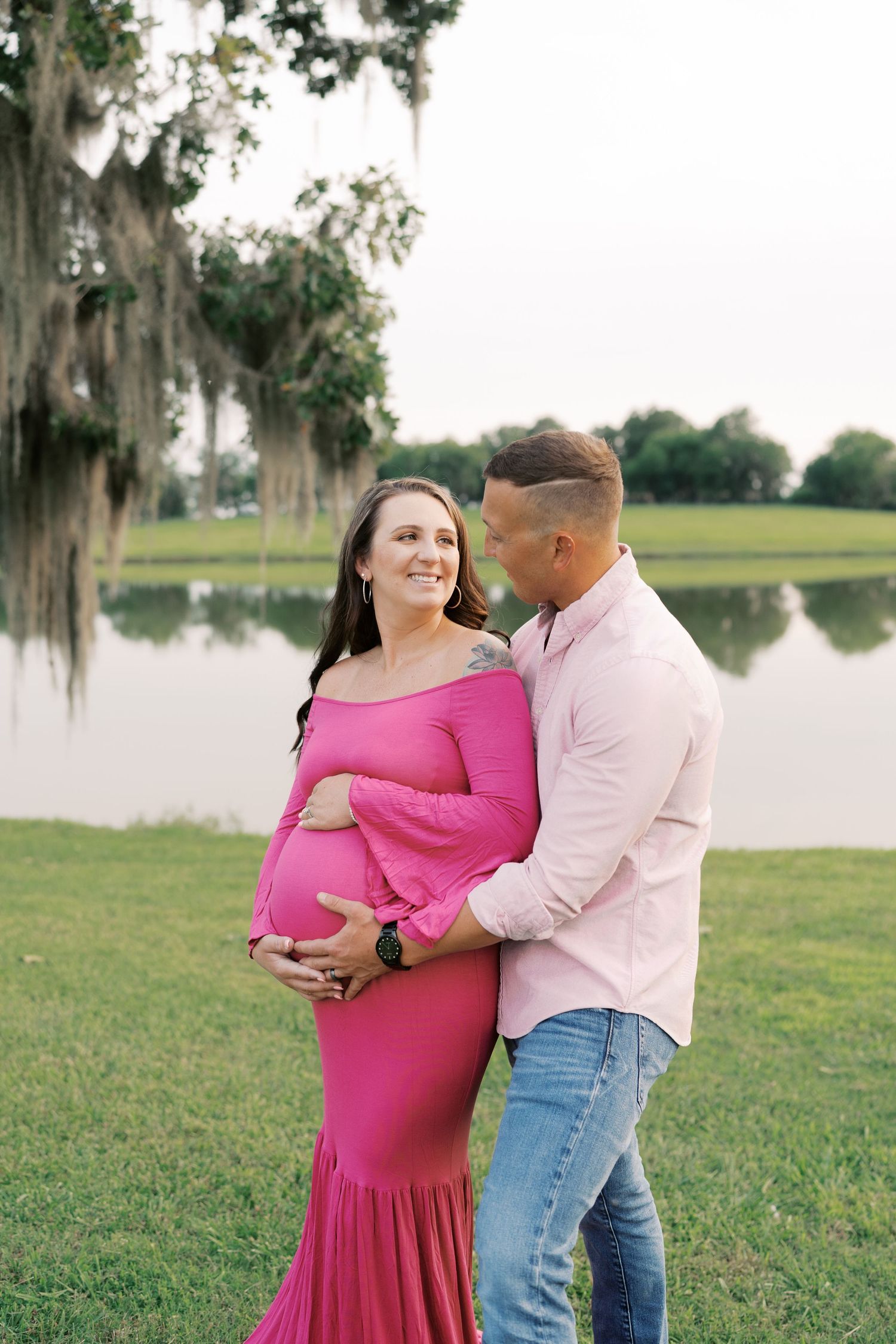 Maternity photos by a lake showing couple embracing under moss-draped trees.