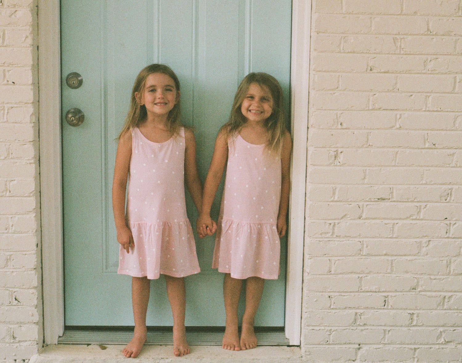 Two children in identical pink dresses stand barefoot in front of a mint green door.