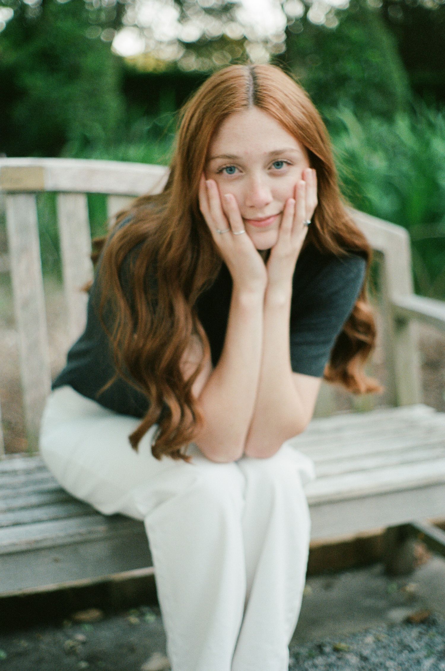 Portrait on wooden bench wearing black top and white pants in garden setting.