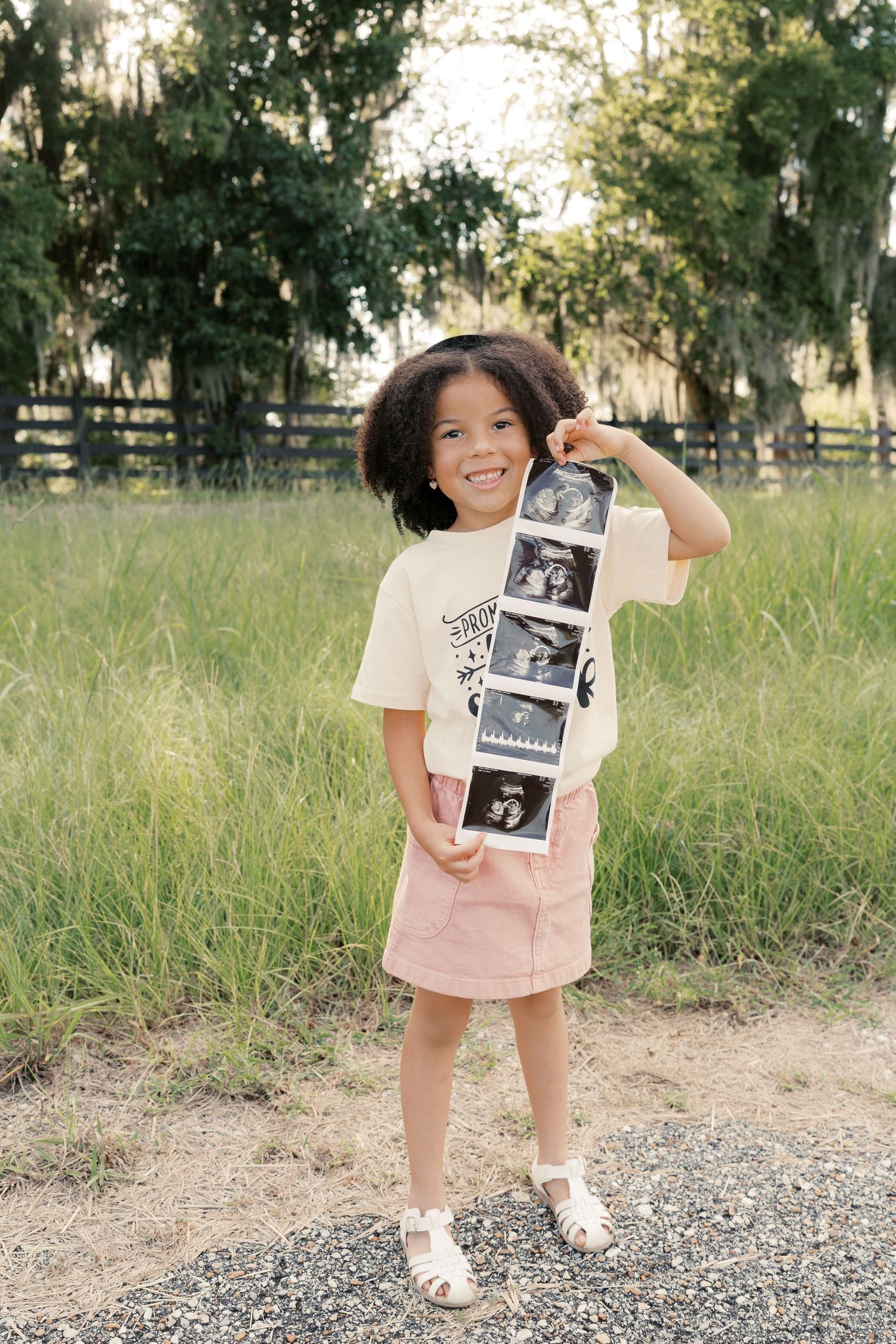 A child in a pink dress holds up numbered ultrasound photos while standing on a gravel path.