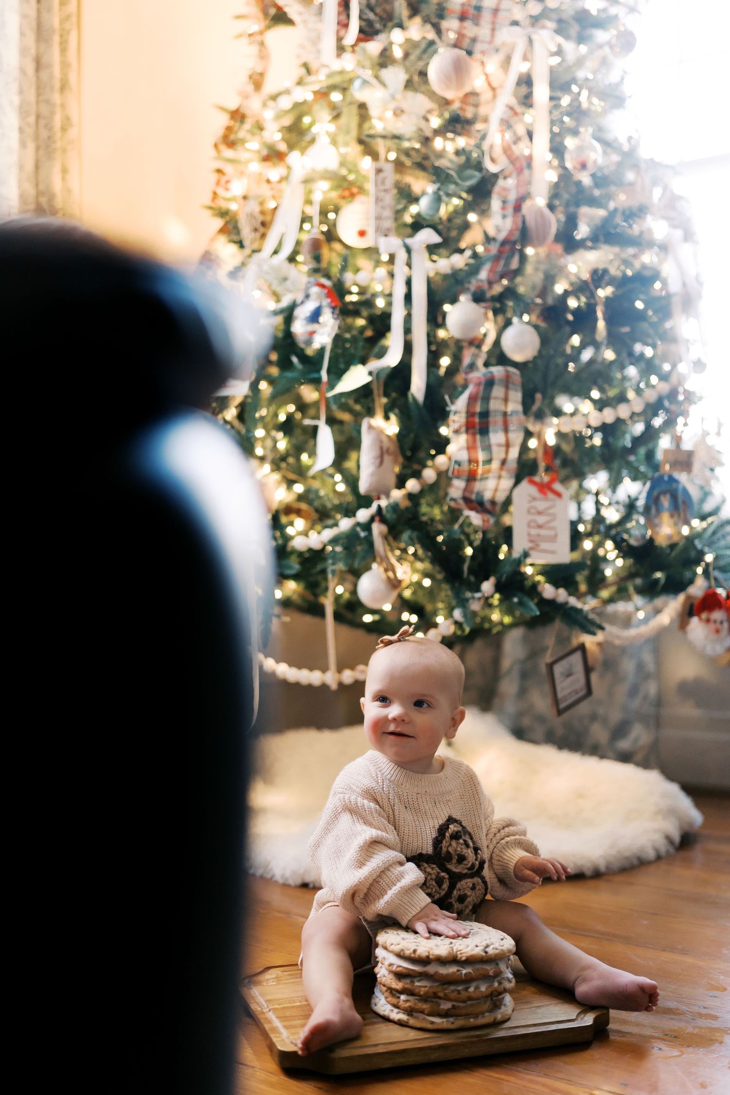 Baby sits on floor with plate of pancakes with Christmas tree twinkling in background.
