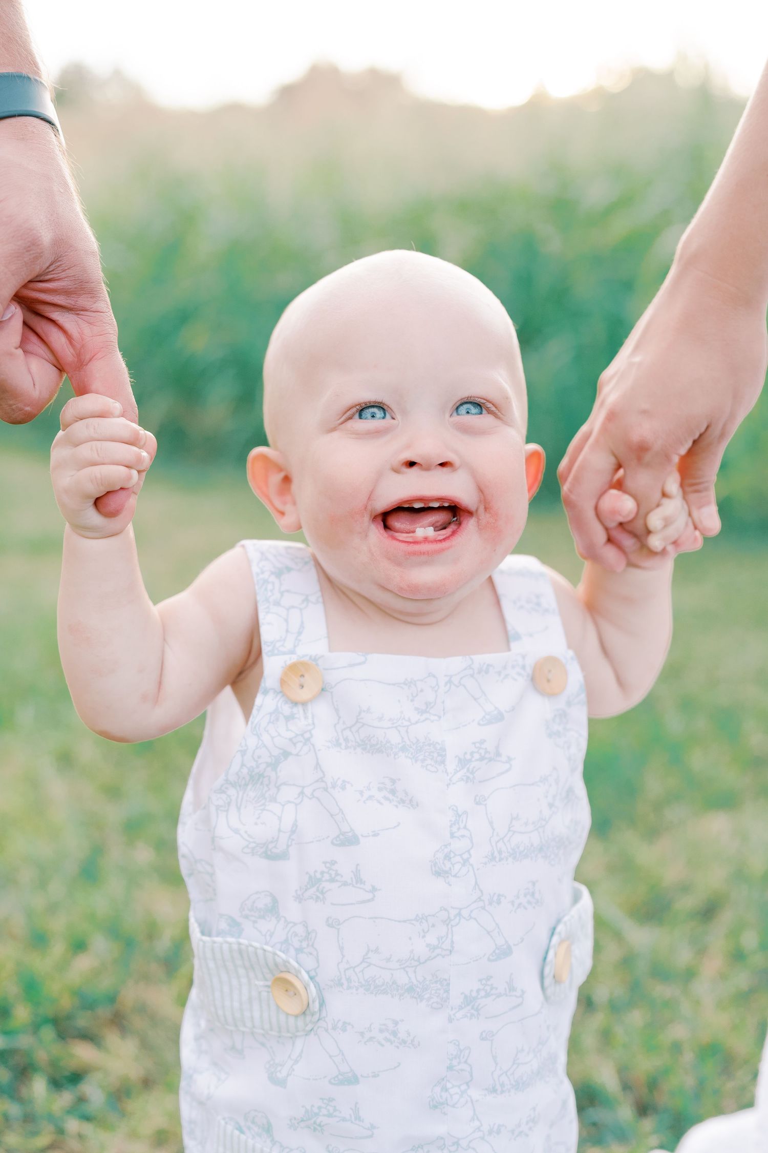 Happy baby smiling and being held up while wearing a white romper in a grassy field.
