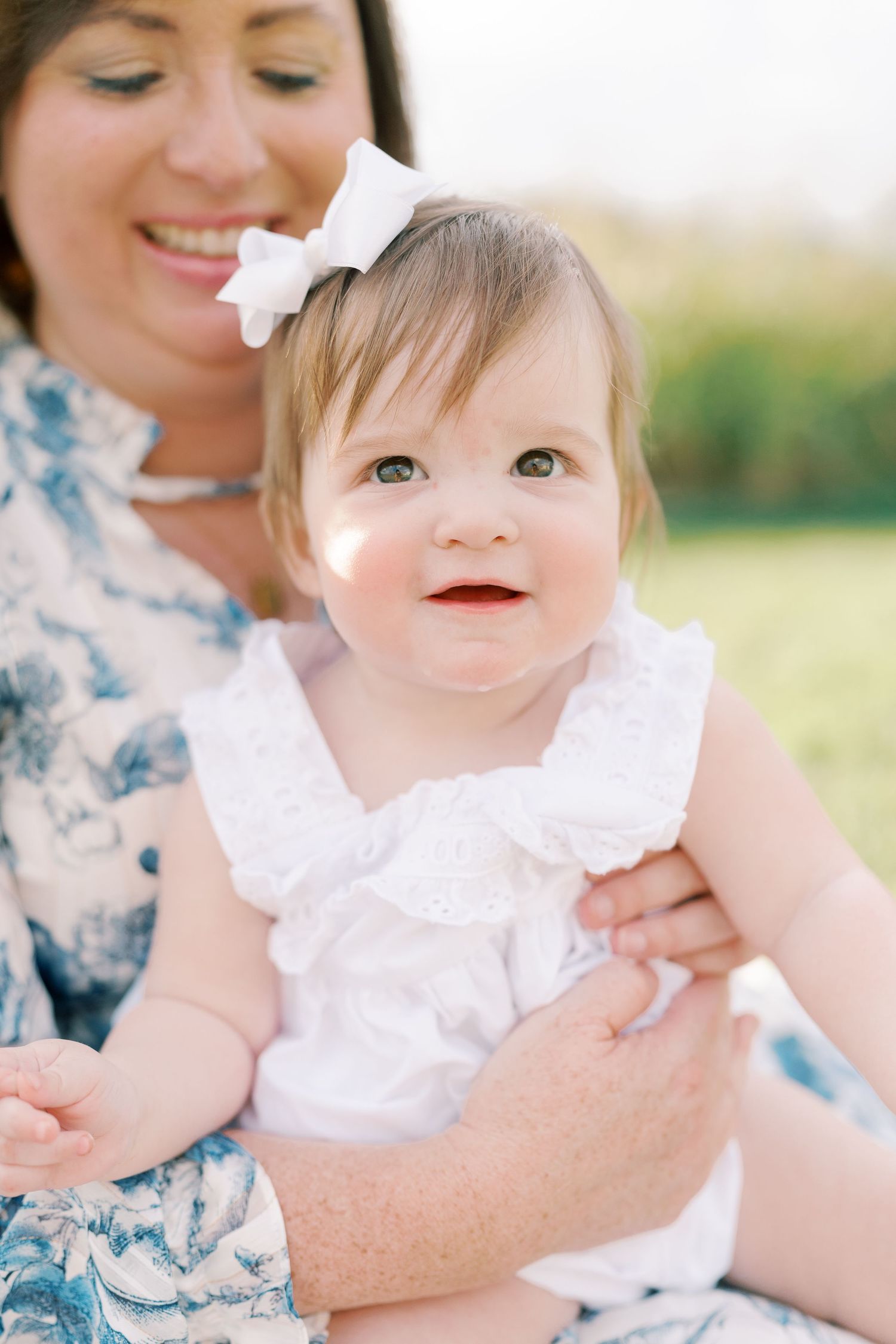 Baby wearing white ruffled dress smiles while being held in a sunlit outdoor setting.