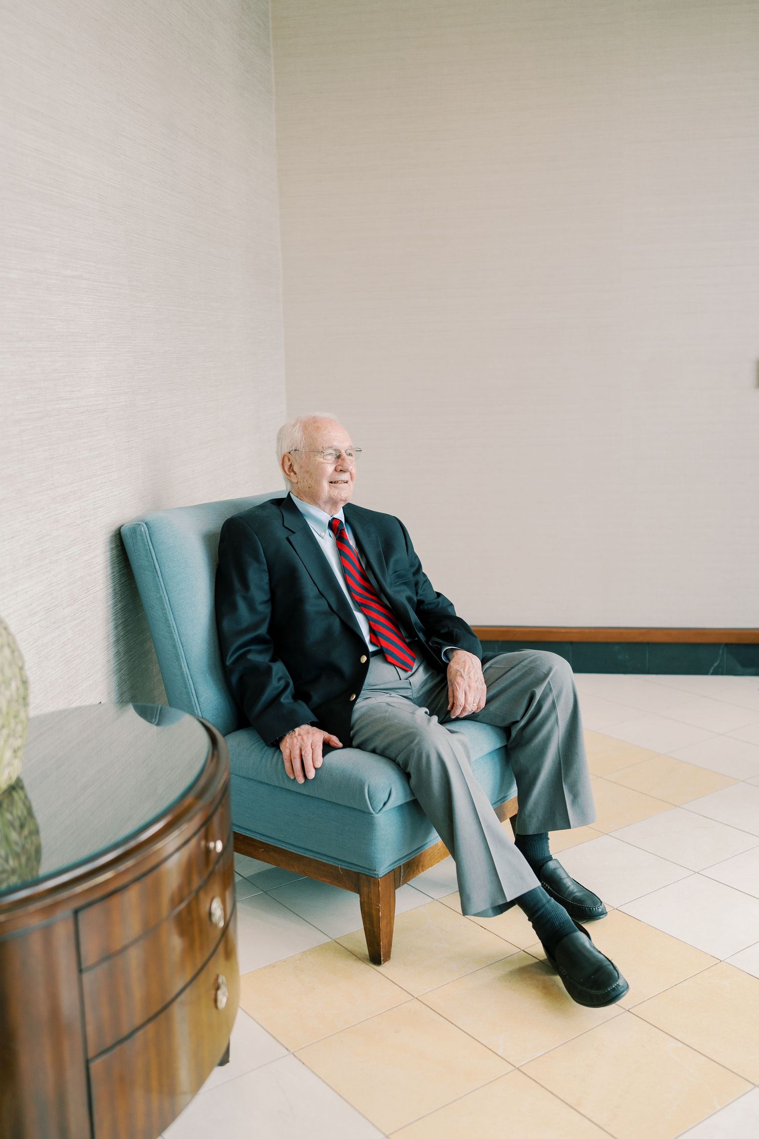 Senior in business attire relaxing on blue armchair against white wall.