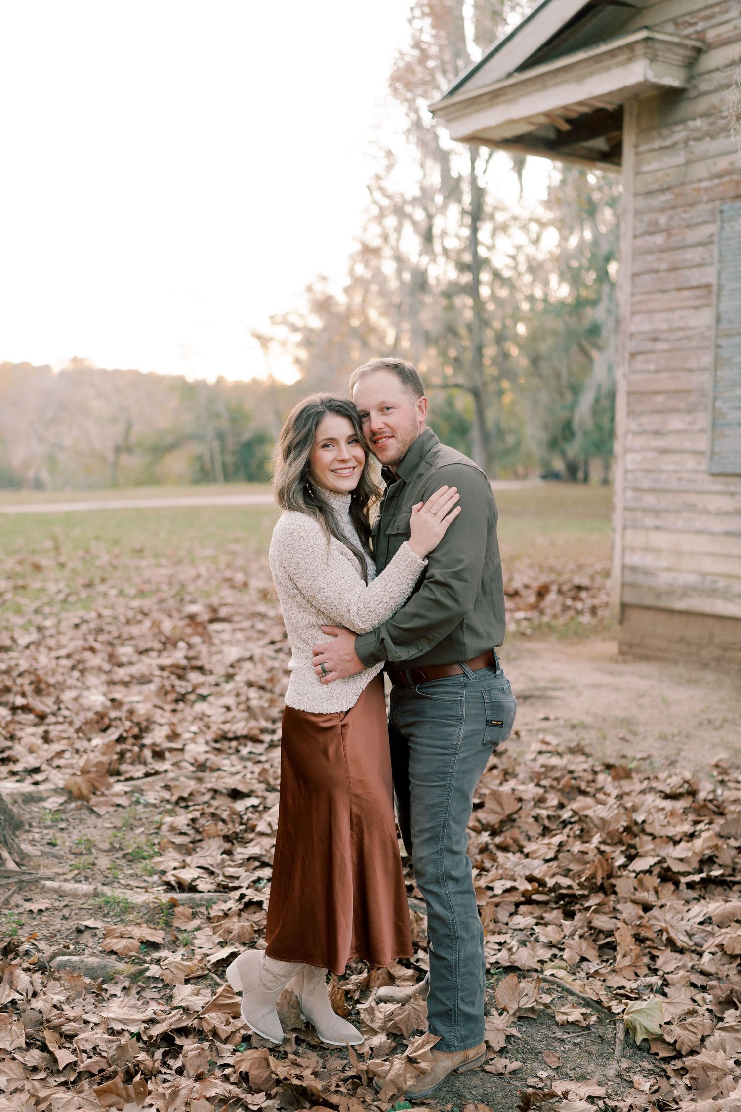 A romantic couple embraces while standing among fallen leaves near a rustic building.