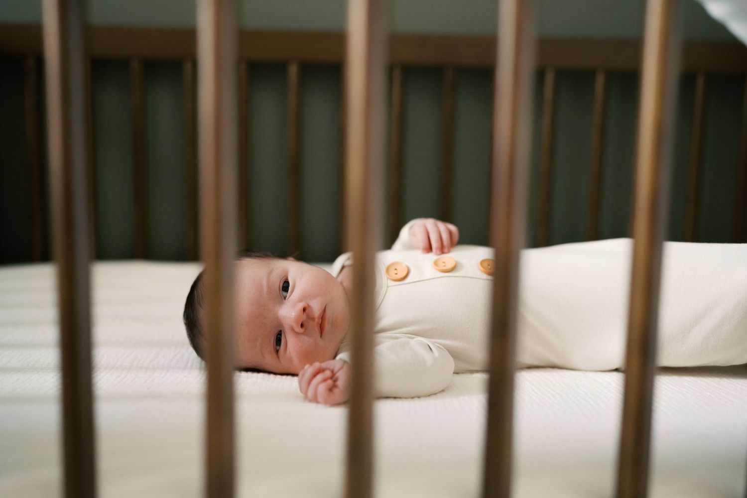 View through crib slats of sleeping newborn in white bedding.