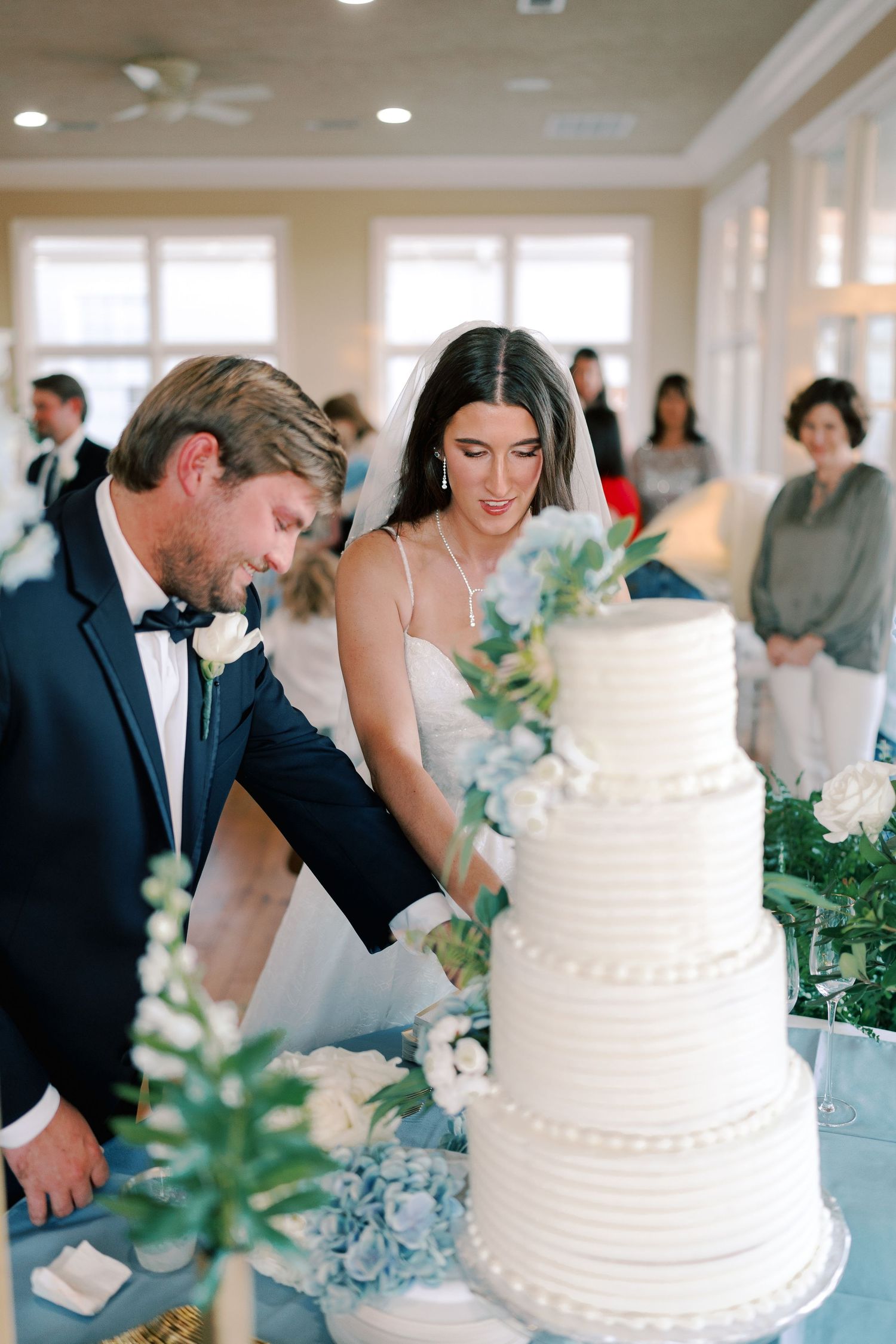 Wedding cake cutting ceremony with elegant white tiered cake decorated with blue flowers.