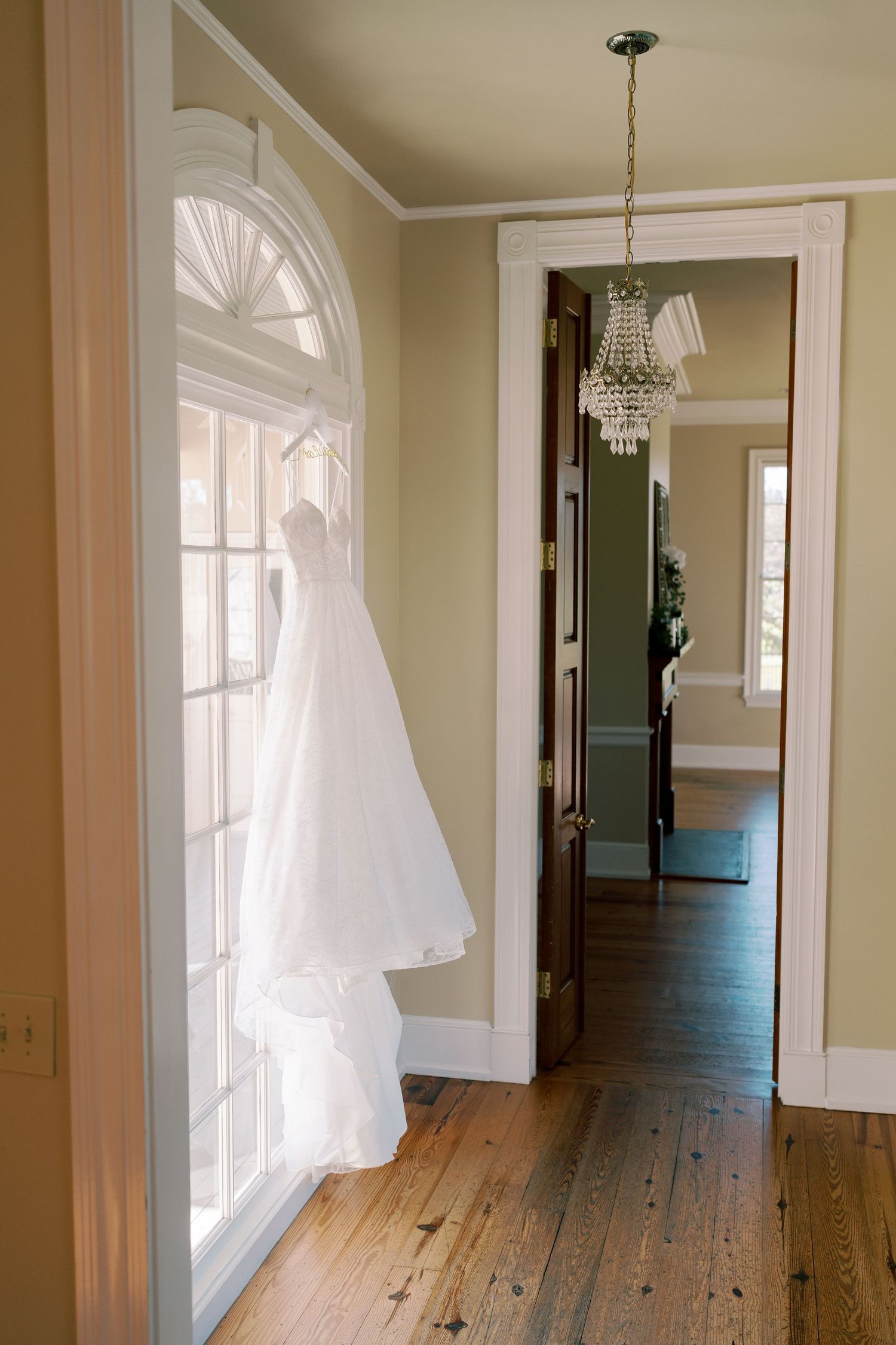 White wedding dress hanging in sunlit doorway with crystal chandelier and hardwood floors.