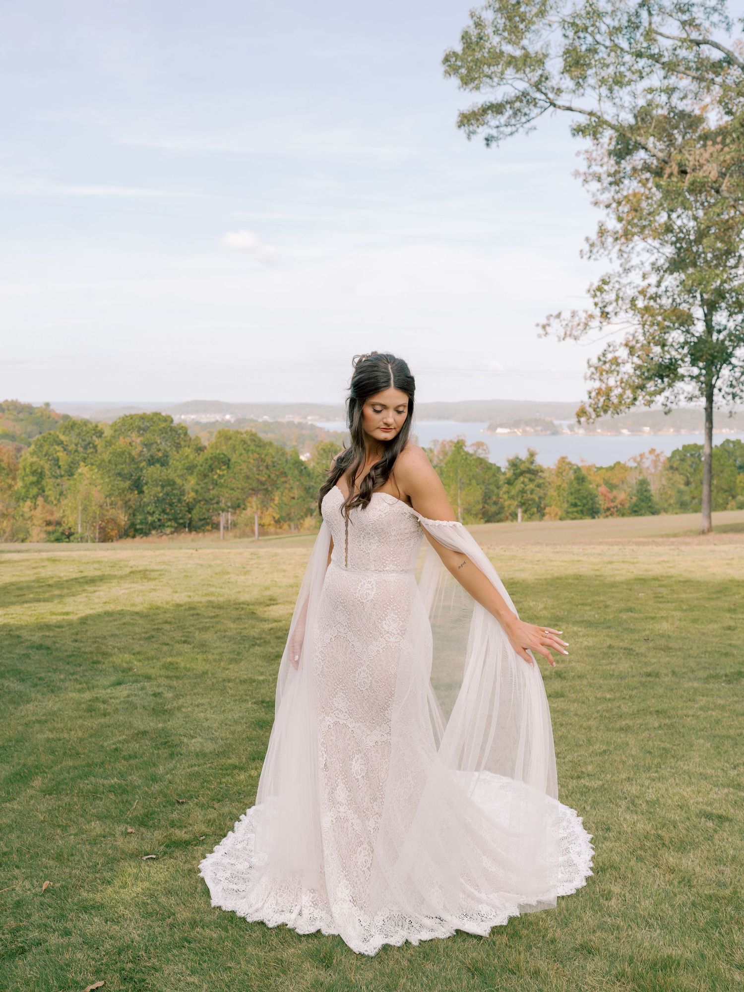 Bride twirls in a lace wedding gown with flowing cape against a scenic mountain backdrop.