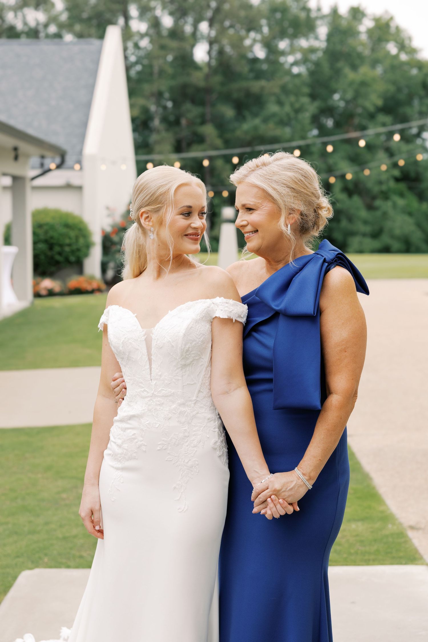 Two women in wedding attire share an emotional embrace outdoors with string lights in the background.
