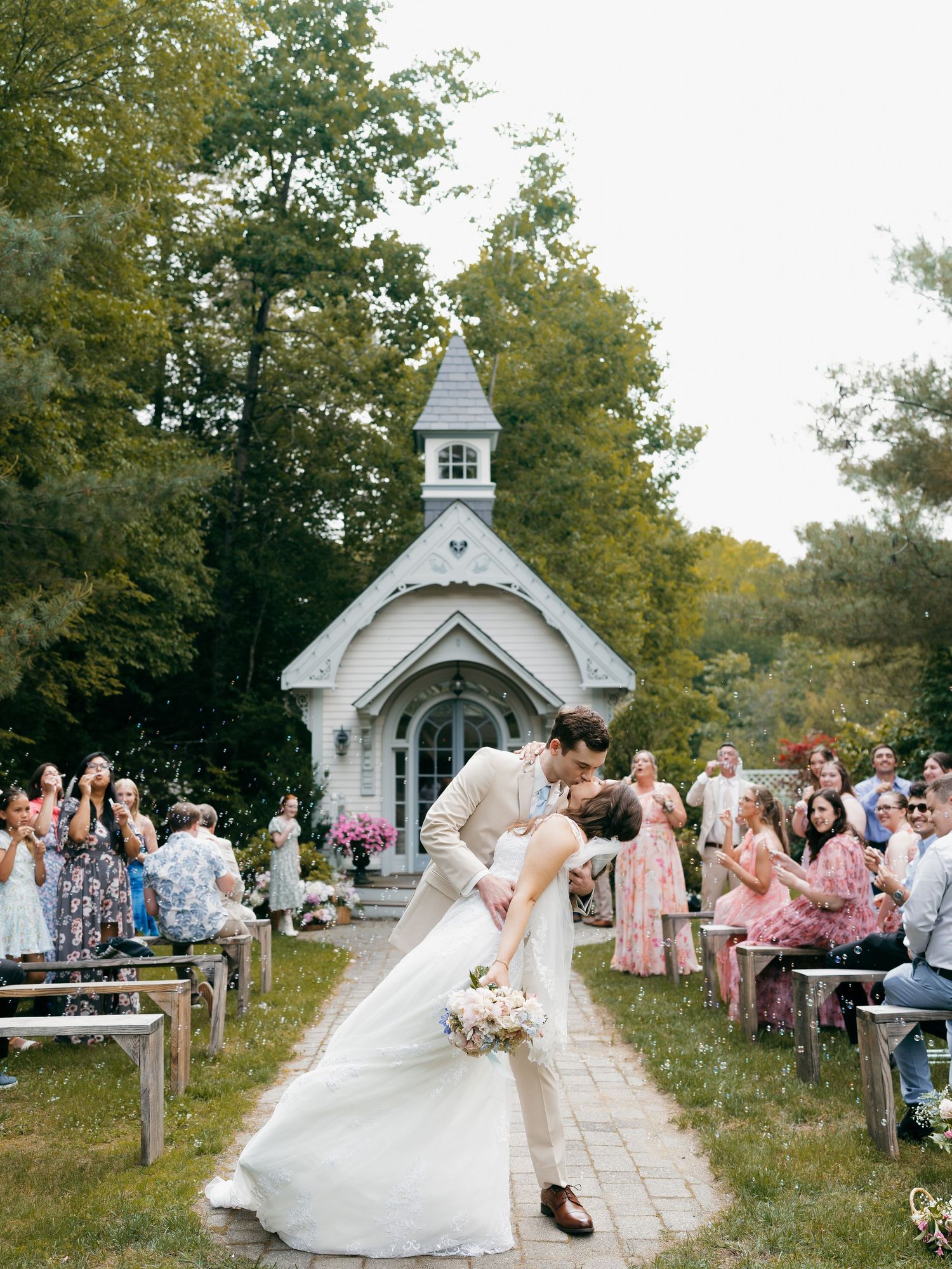 Wedding ceremony outside charming white chapel with guests seated along pathway.
