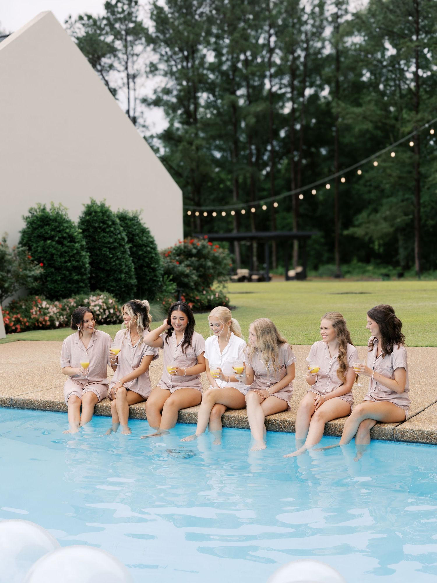 Wedding party sitting at pool edge with feet in water during outdoor celebration.