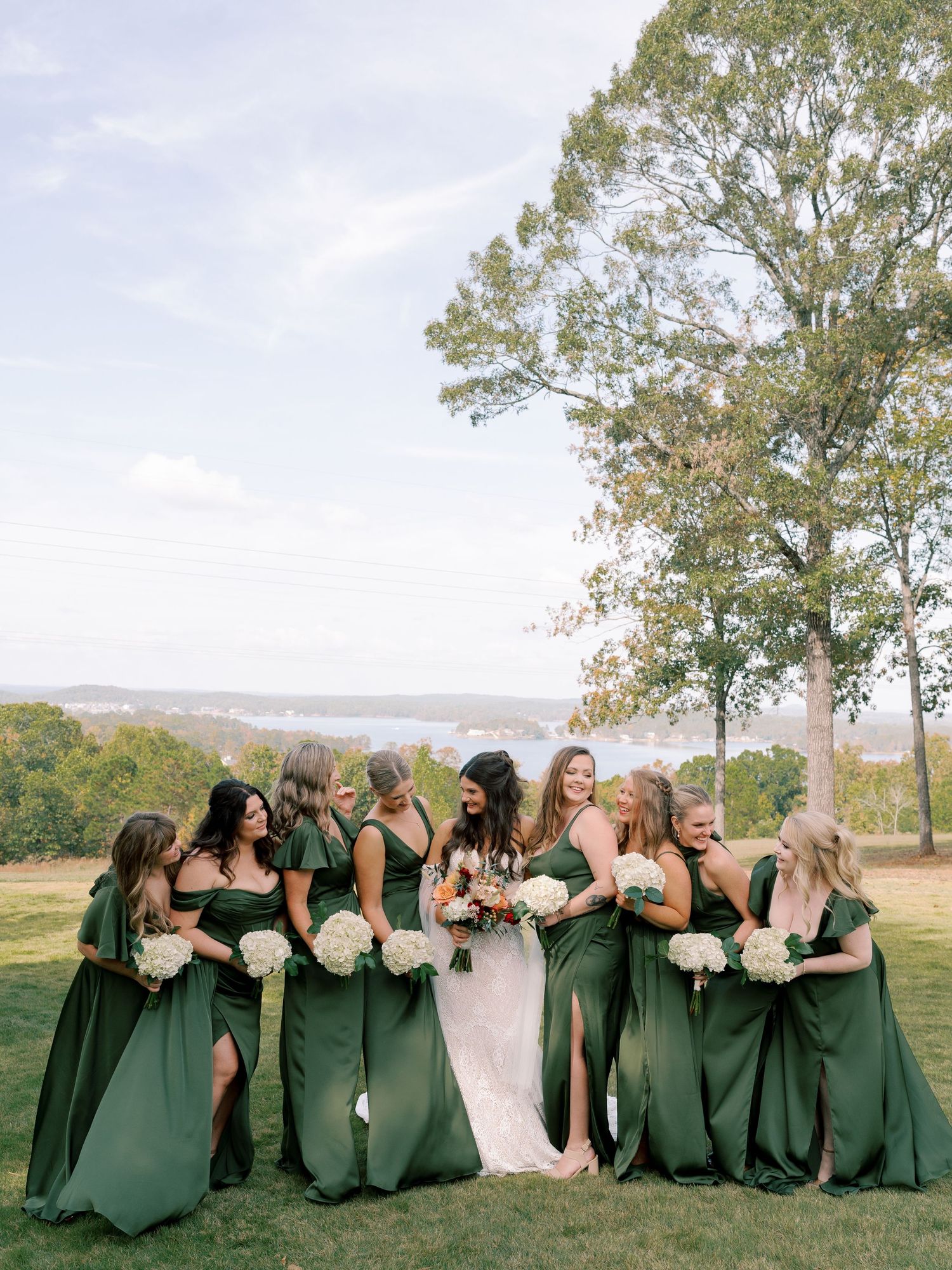 Bridal party in dark green dresses creates fun photo moments on a scenic hilltop.