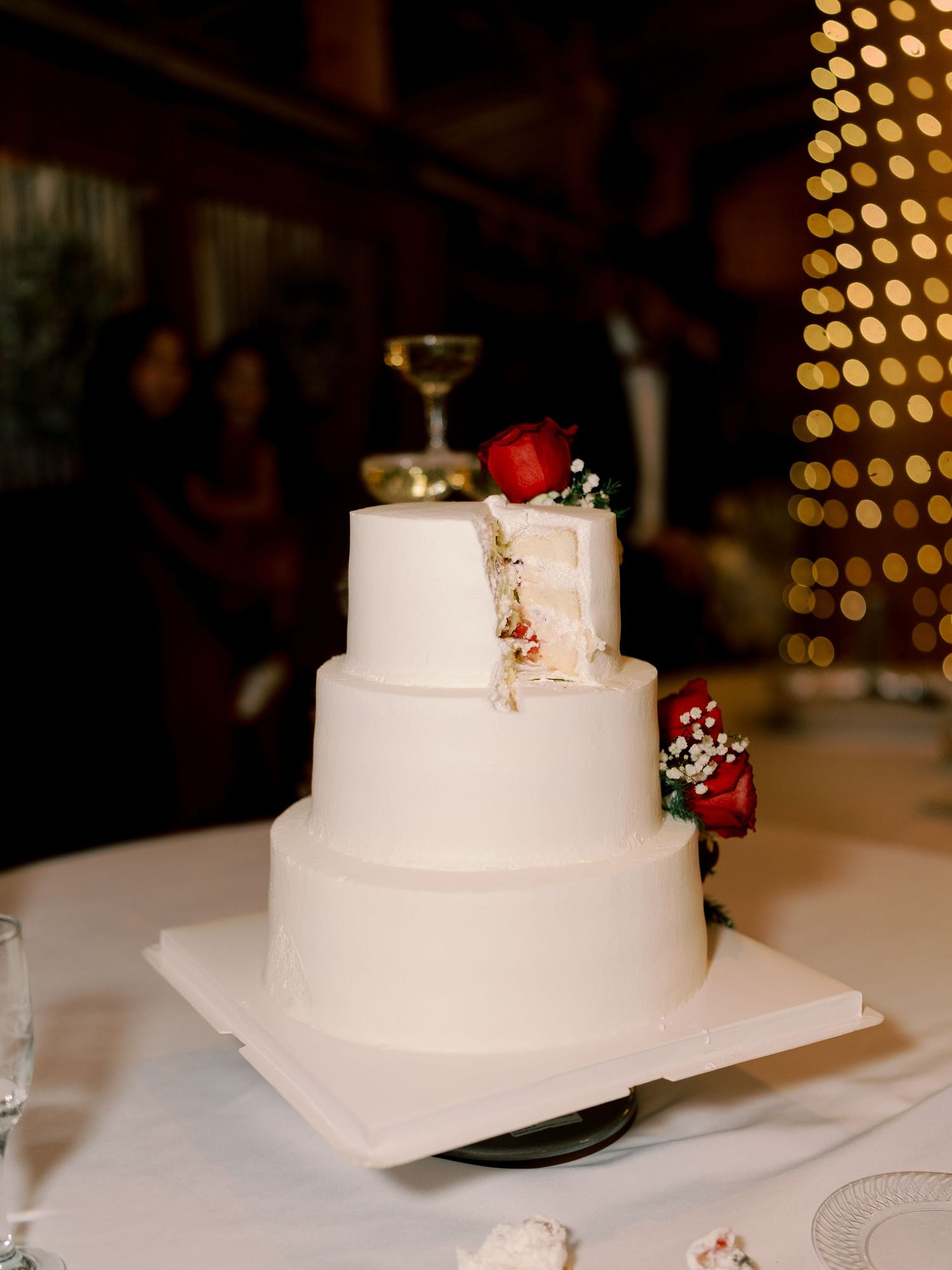 White tiered wedding cake with red rose decorations displayed at reception.