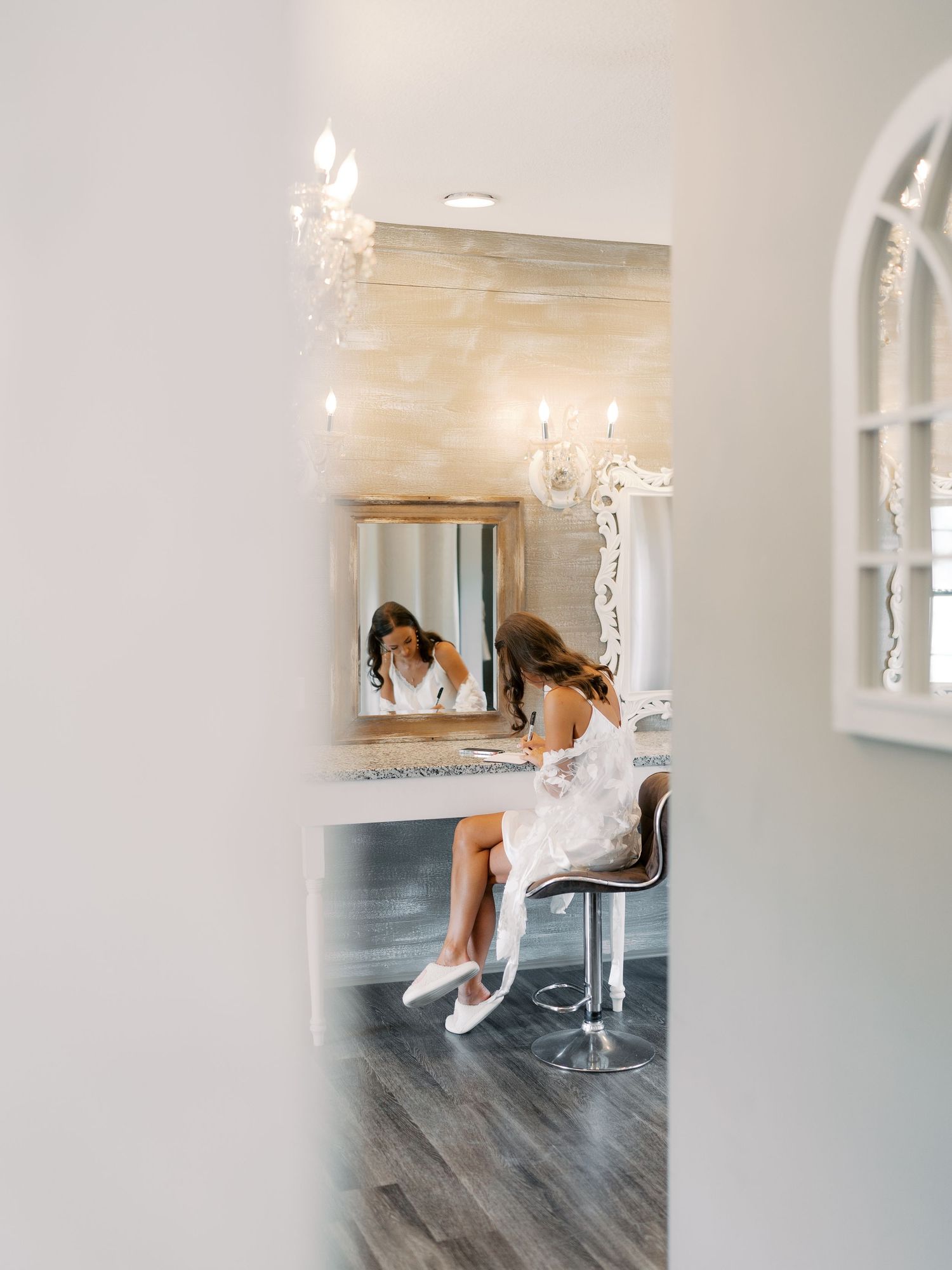 A wedding dress hangs elegantly in a bright white room with a crystal chandelier and grey flooring.