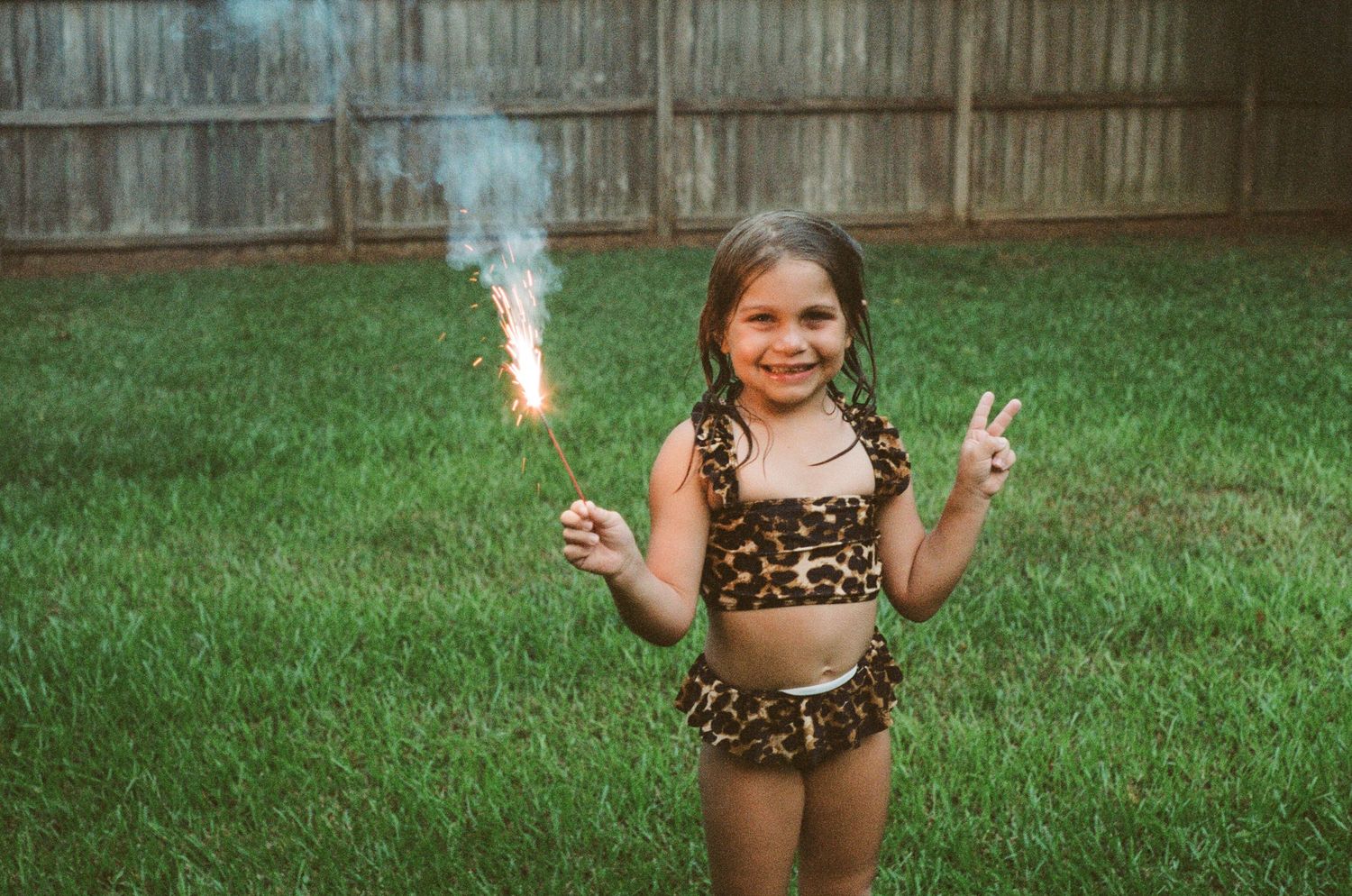Child holding sparklers and playing outdoors by a wooden fence at dusk.