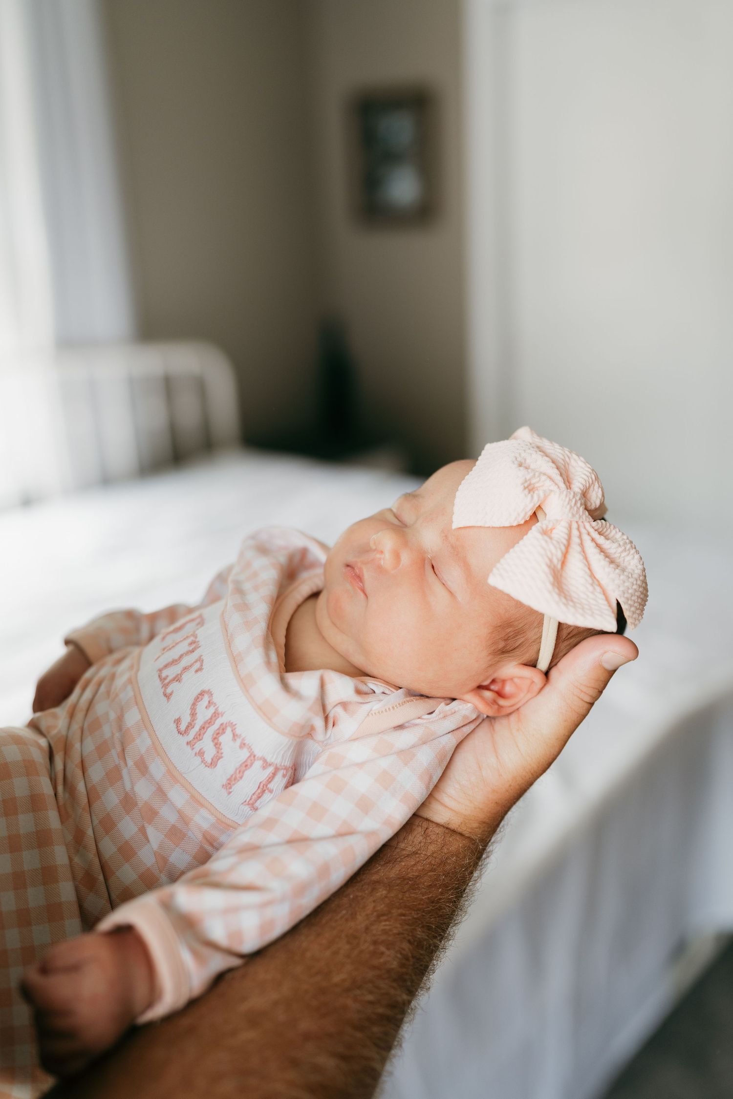 Newborn baby wearing pink gingham sleeper rests peacefully on parent's arm.