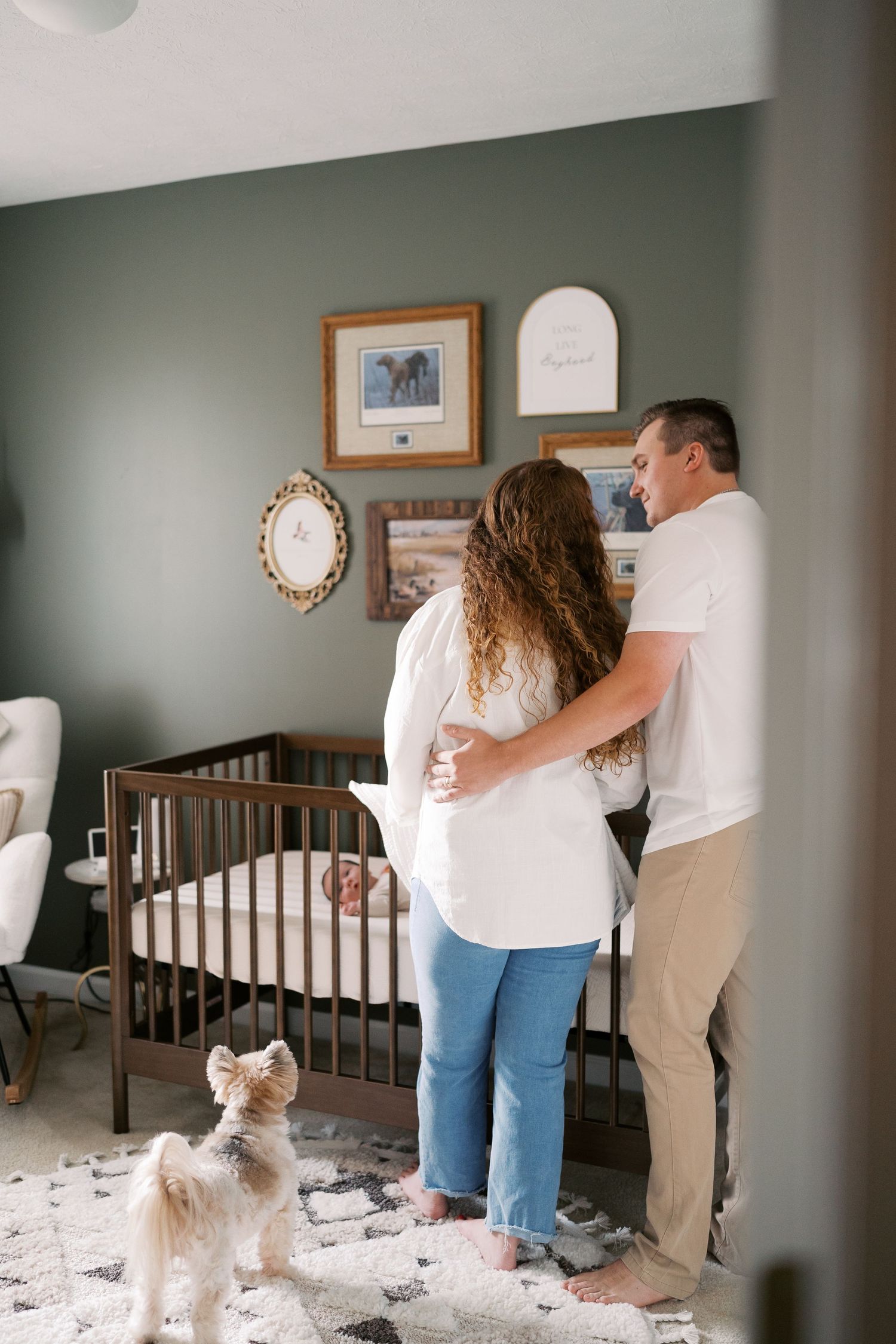 A couple stands together in a cozy nursery room with sage green walls and white furniture.