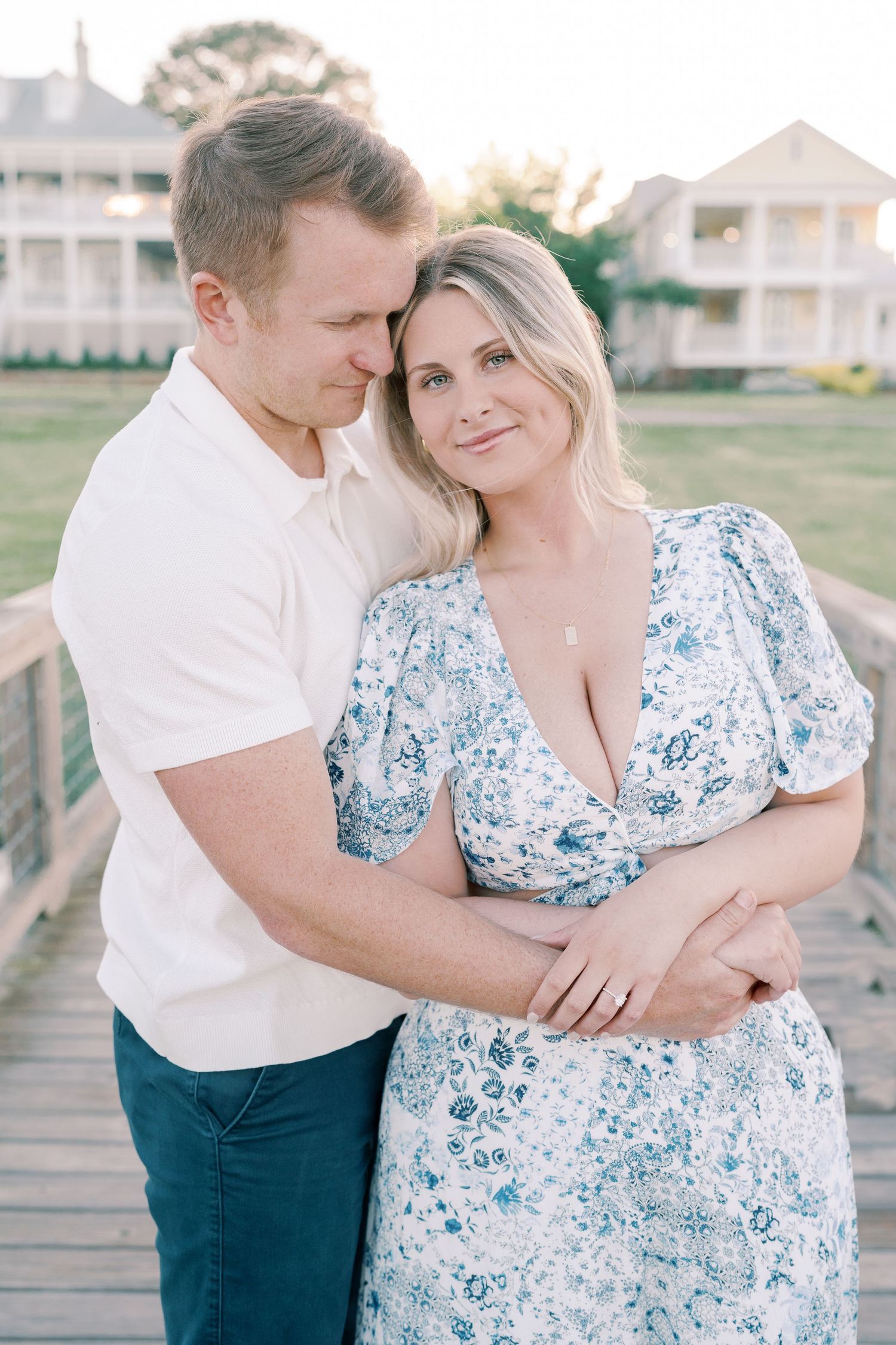 Romantic couple embraces on boardwalk with coastal homes in background.