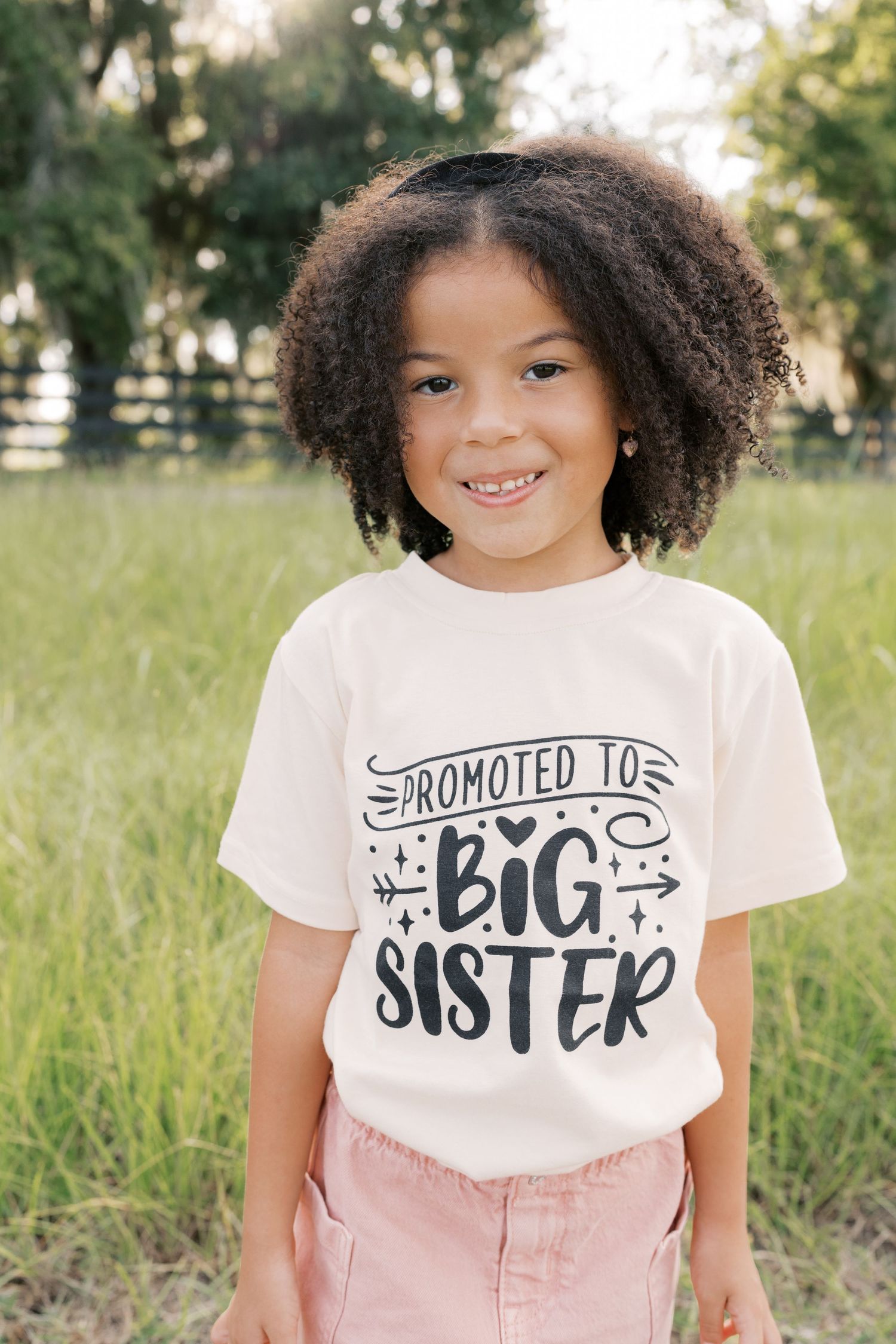 A child wearing a 'Promoted to Big Sister' t-shirt smiles brightly in an outdoor setting.