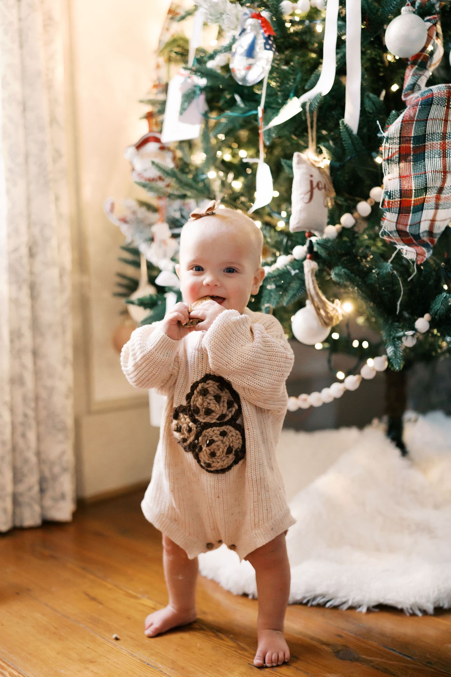 Baby wearing knit sweater crawls near Christmas tree on hardwood floor.