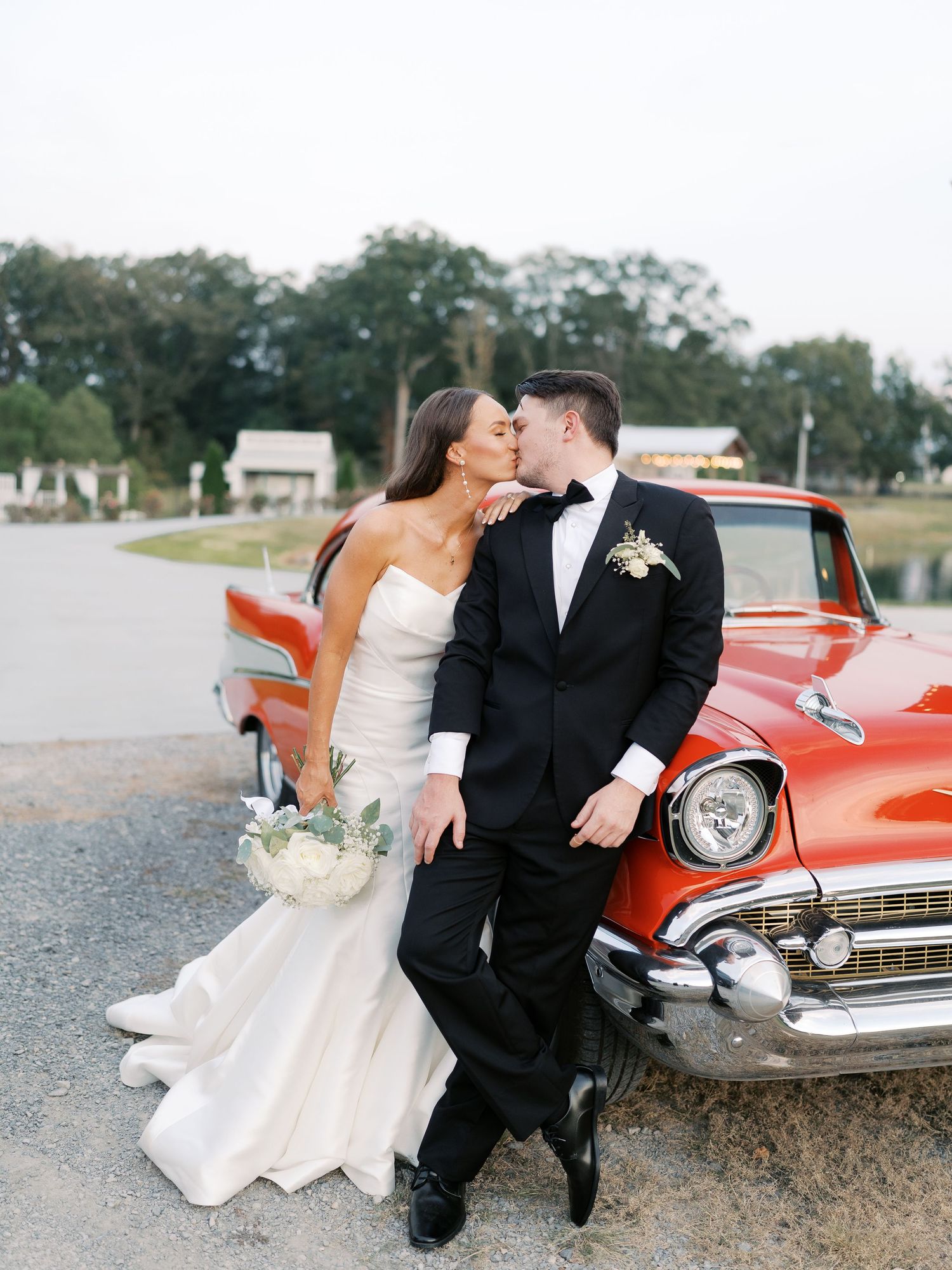 Couple in wedding attire posing by vintage red car at sunset.
