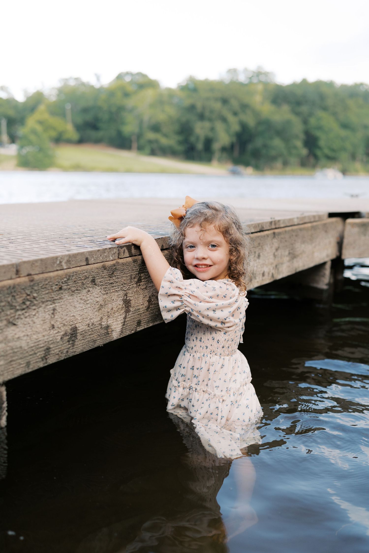 A child in a floral dress plays at the edge of a wooden dock over calm water.