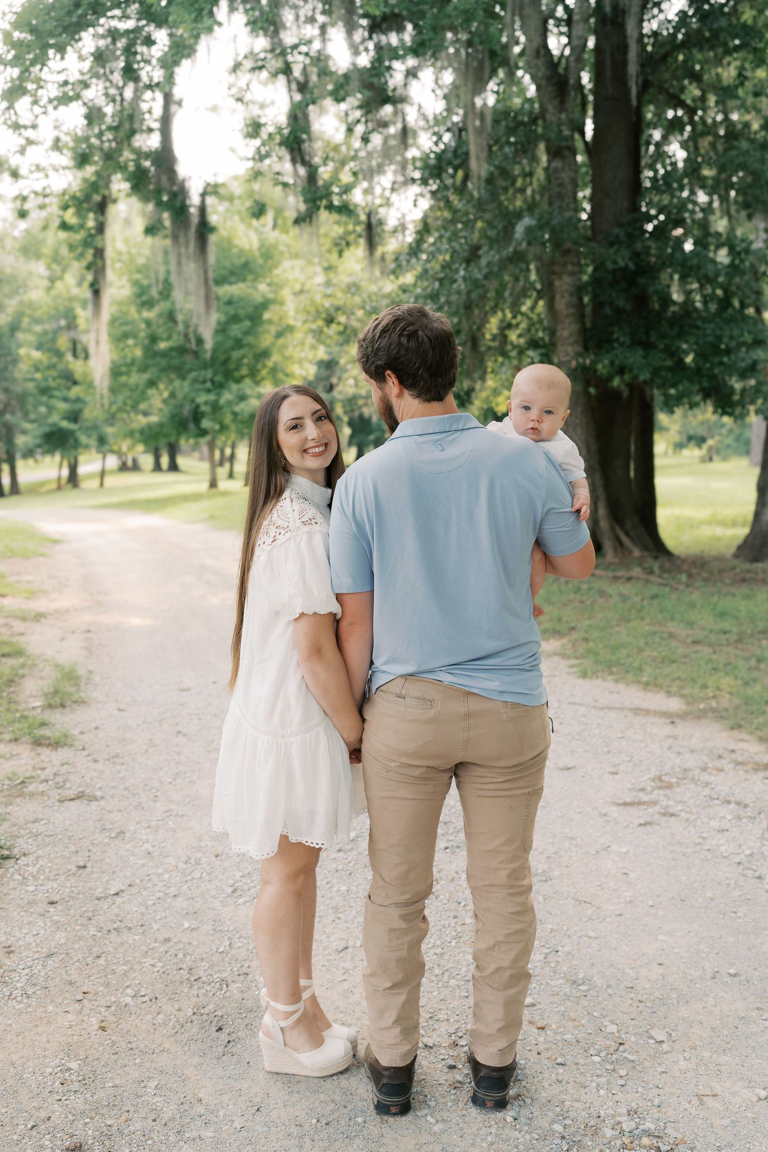 A family walks together on a tree-lined dirt path in a park on a sunny day.