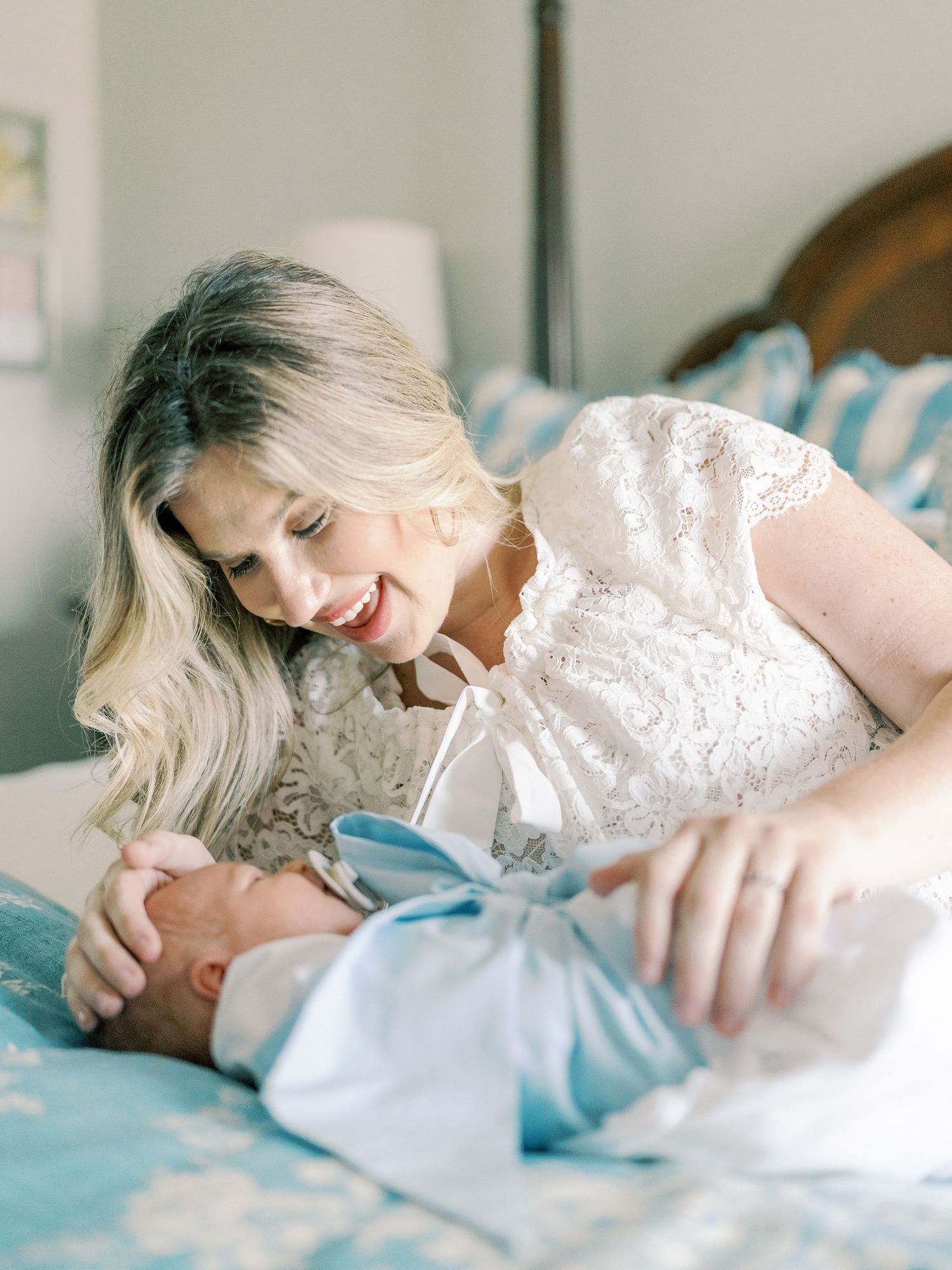 Mother in white lace dress cuddles newborn on blue floral bedding.
