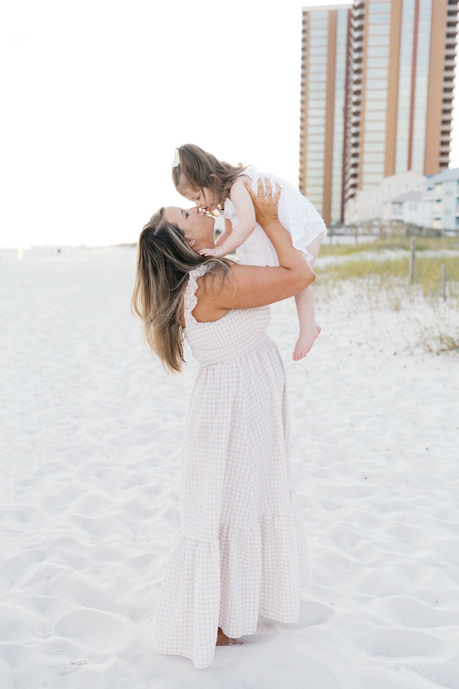 A parent and child share joyful moments together on a white sandy beach with condos in background.