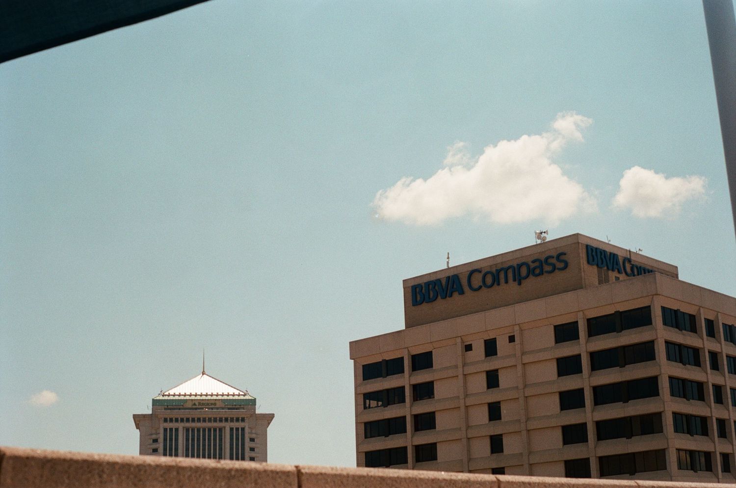 Modern office buildings and skyscrapers rise against a blue sky with white clouds.