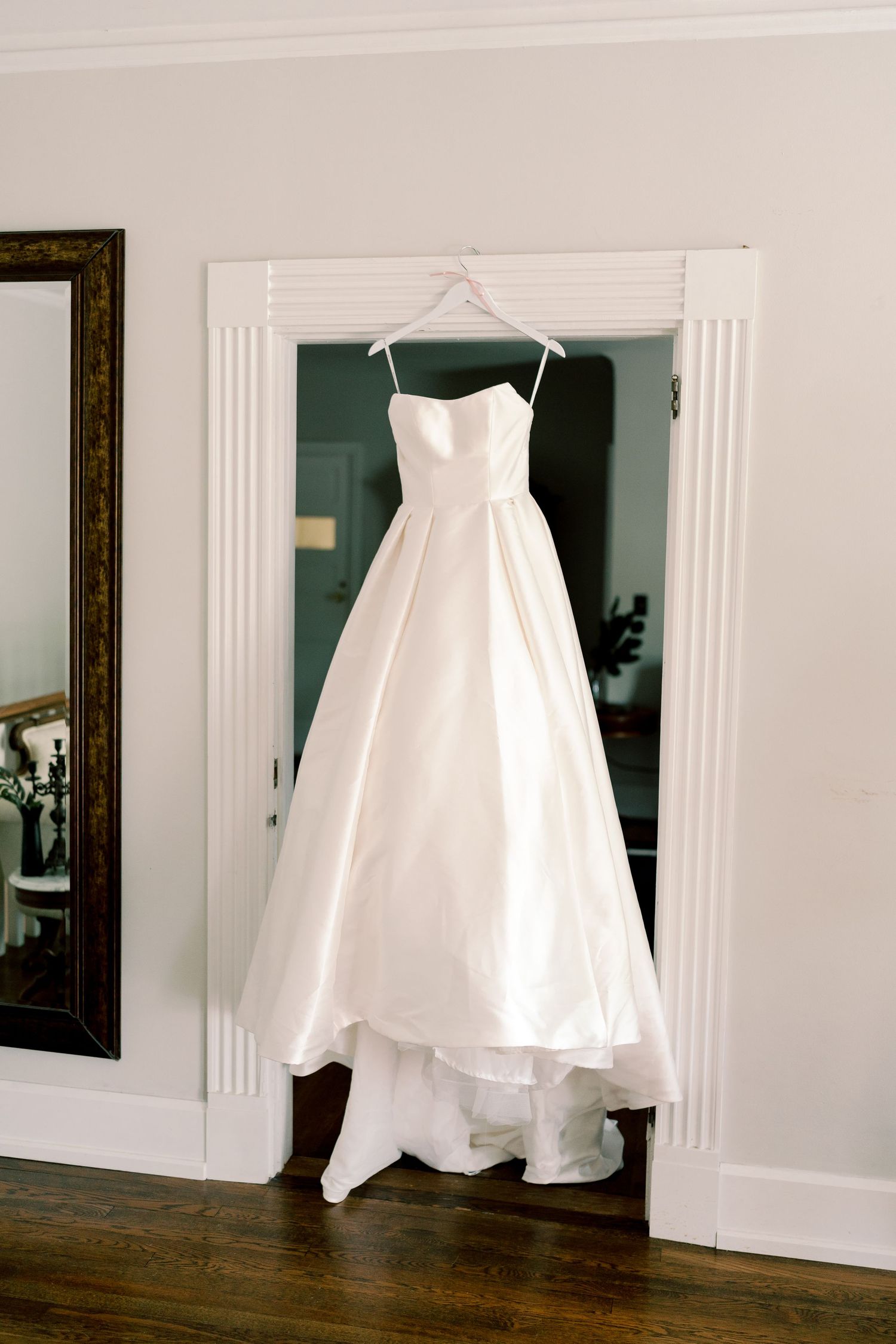 A white wedding dress hangs elegantly against a light-colored wall with decorative molding.