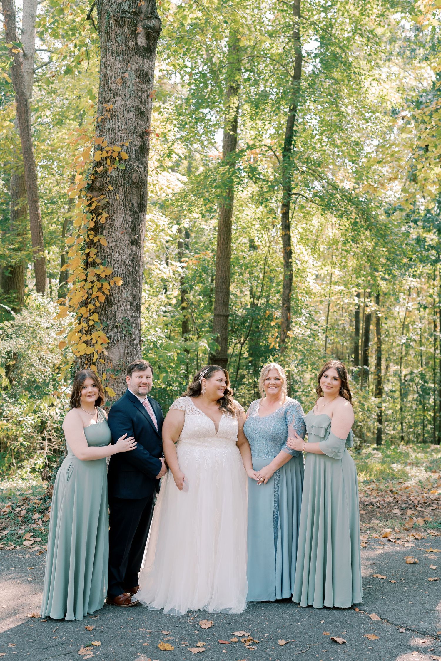 Wedding party in sage green dresses and black suit laughing together in a wooded setting.
