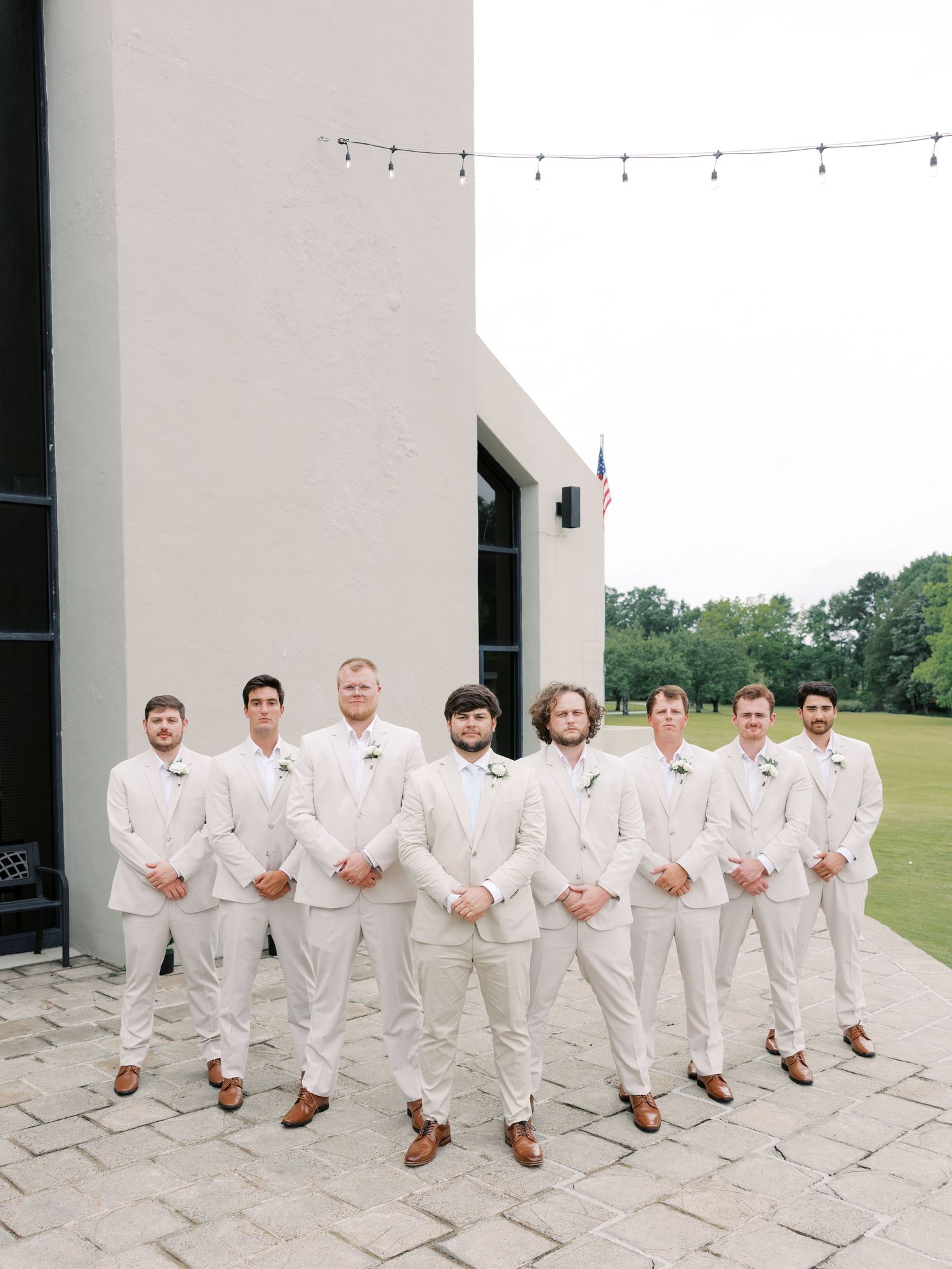 Groomsmen in crisp white suits and brown shoes pose on a stone patio.