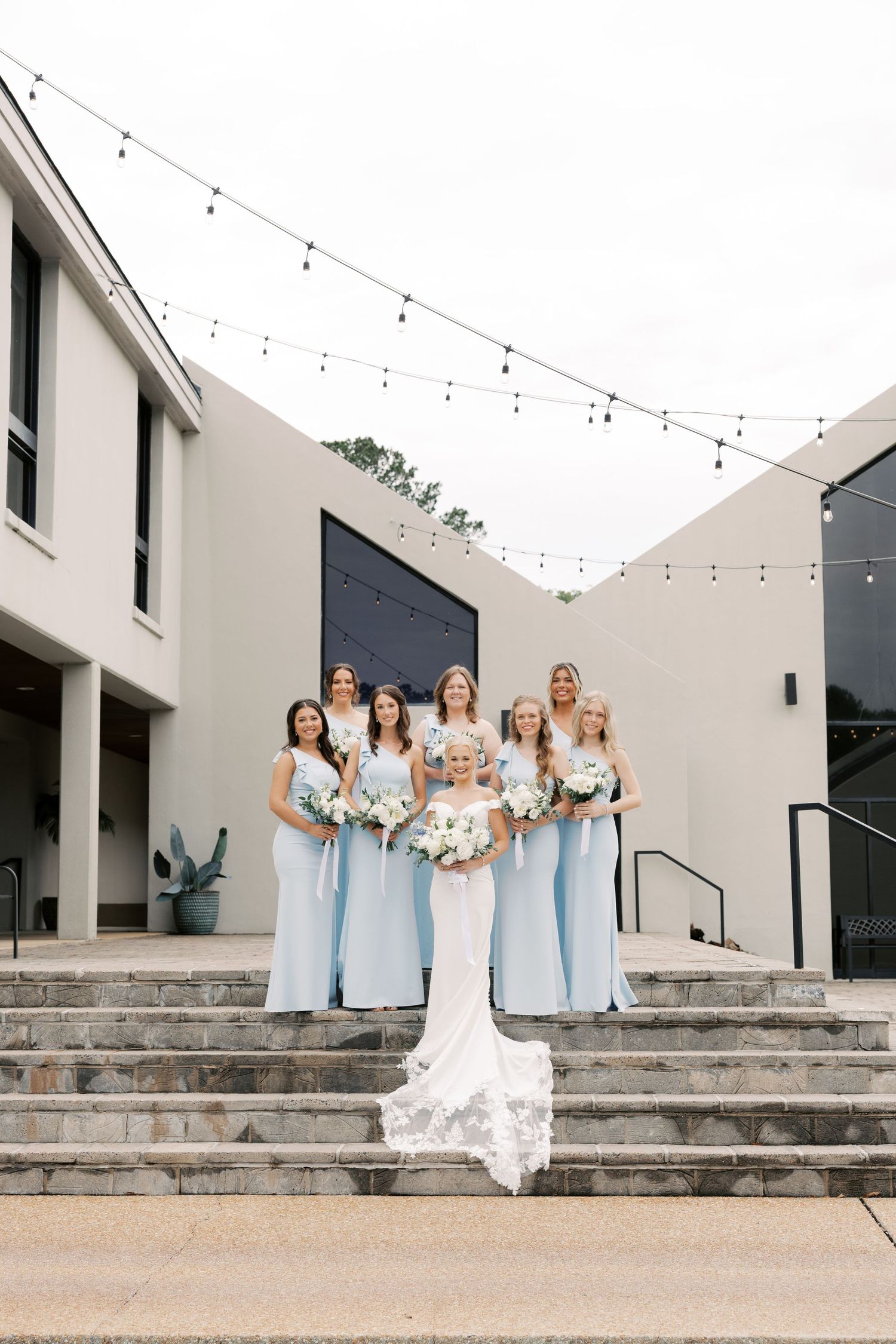 Bridal party in powder blue dresses gather on modern architectural steps for formal photos.