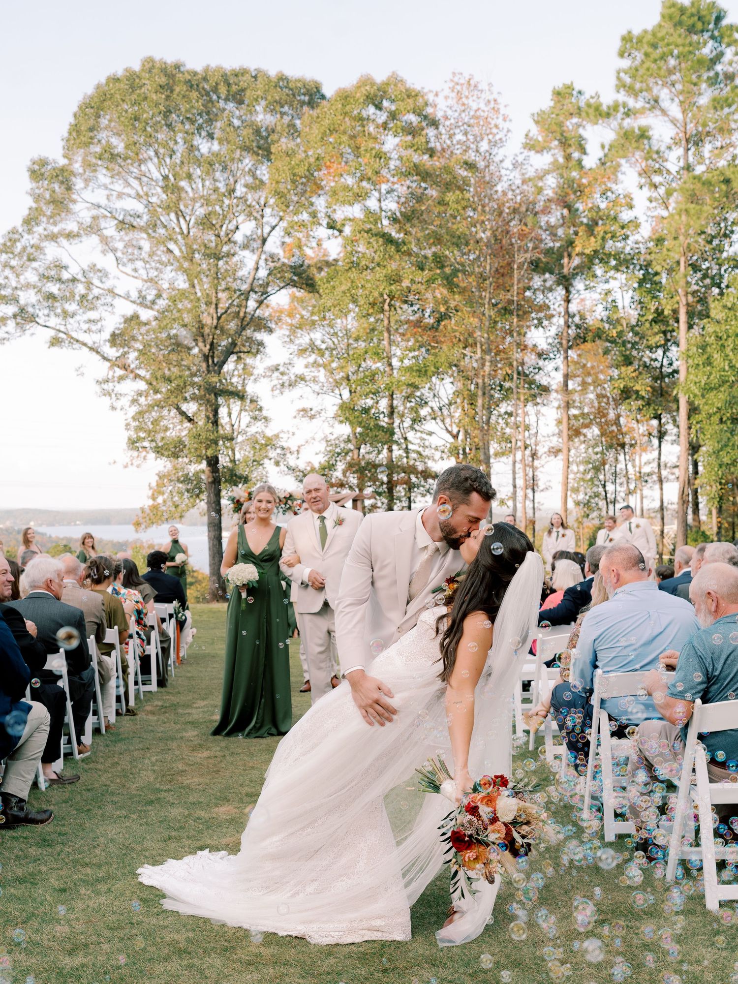 Wedding guests seated on white chairs watching outdoor ceremony with pine trees in background.
