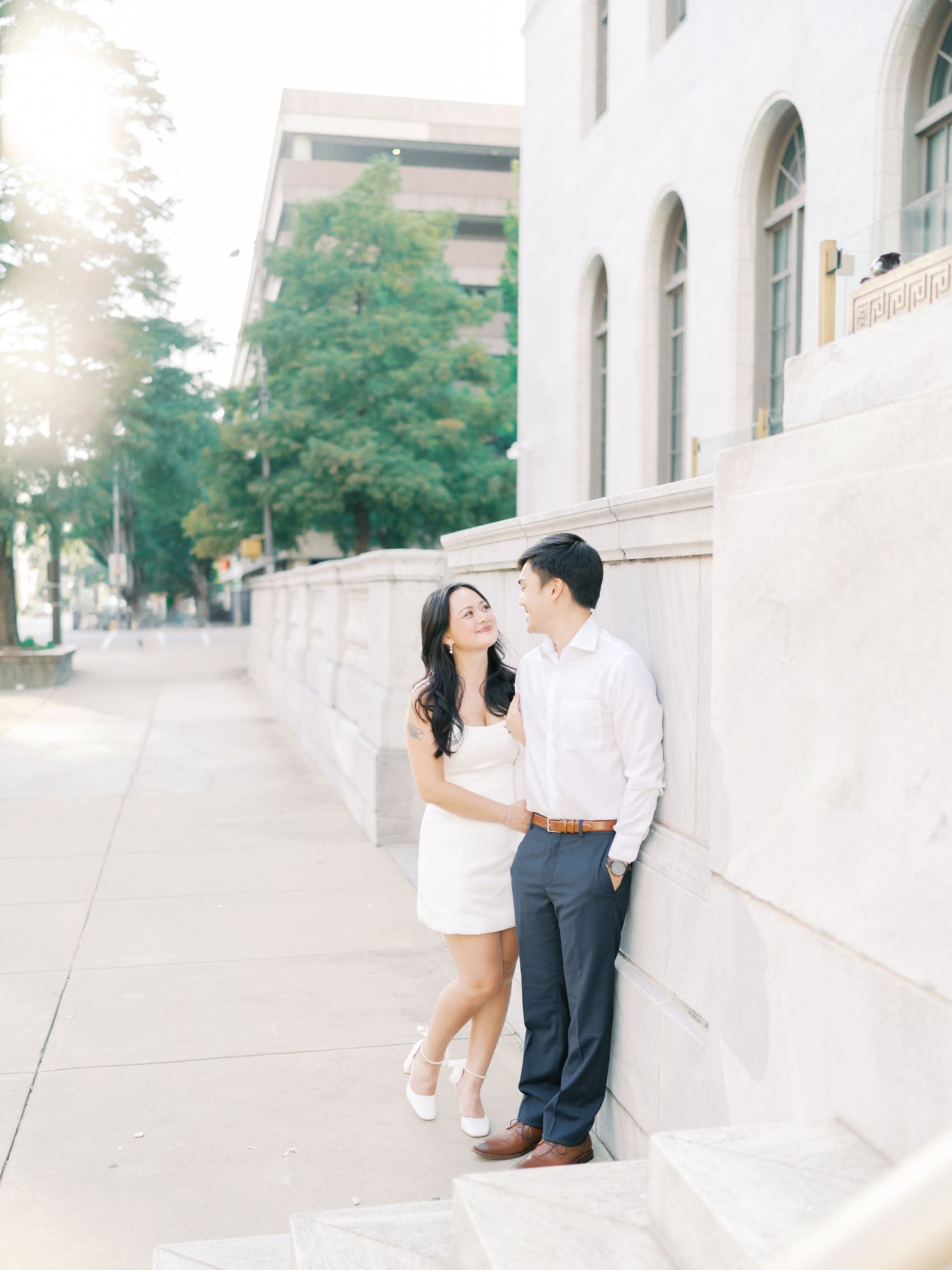 A couple shares intimate moments against a white building wall during a sunny outdoor photoshoot.