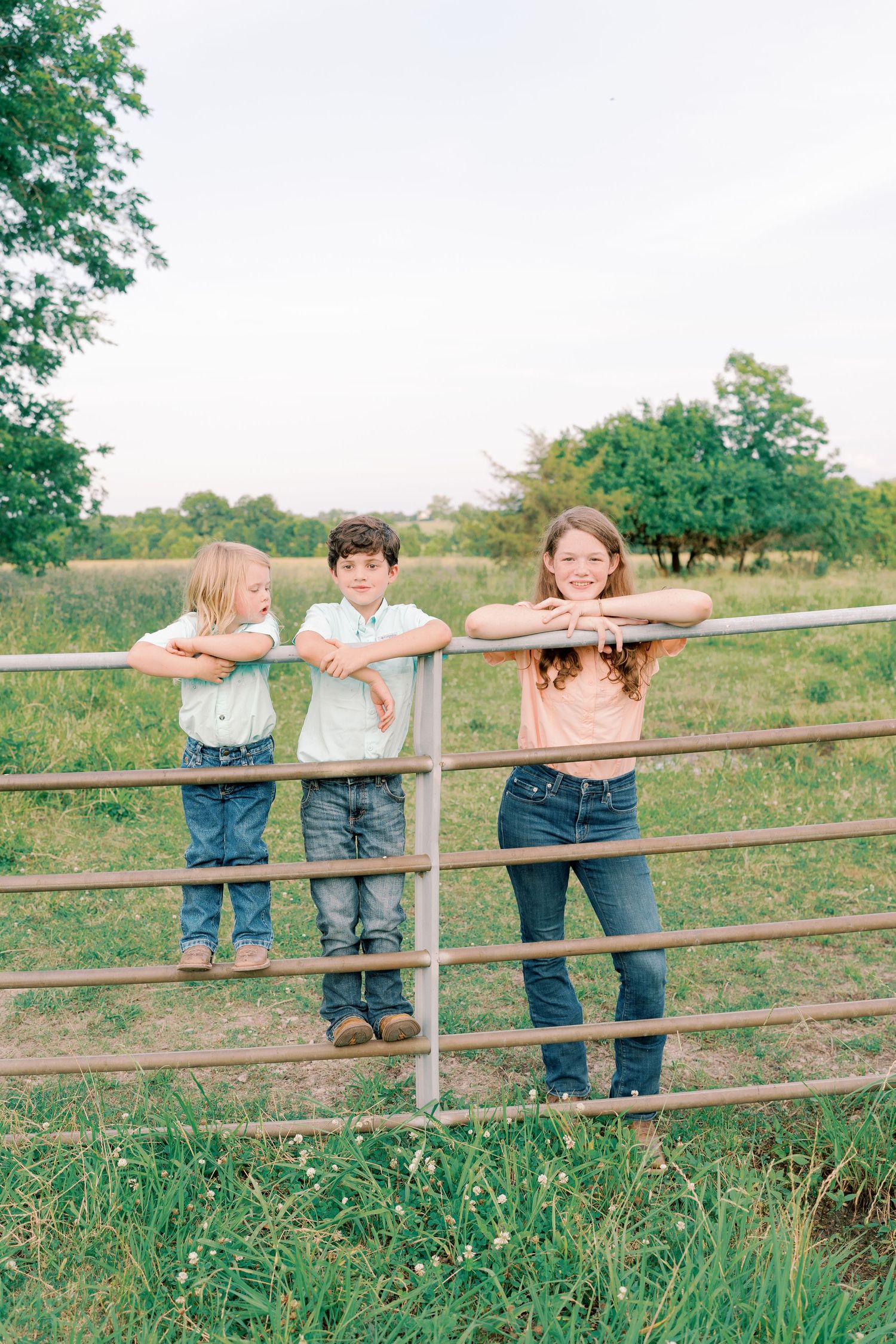Three children lean on metal gate rails in countryside setting.