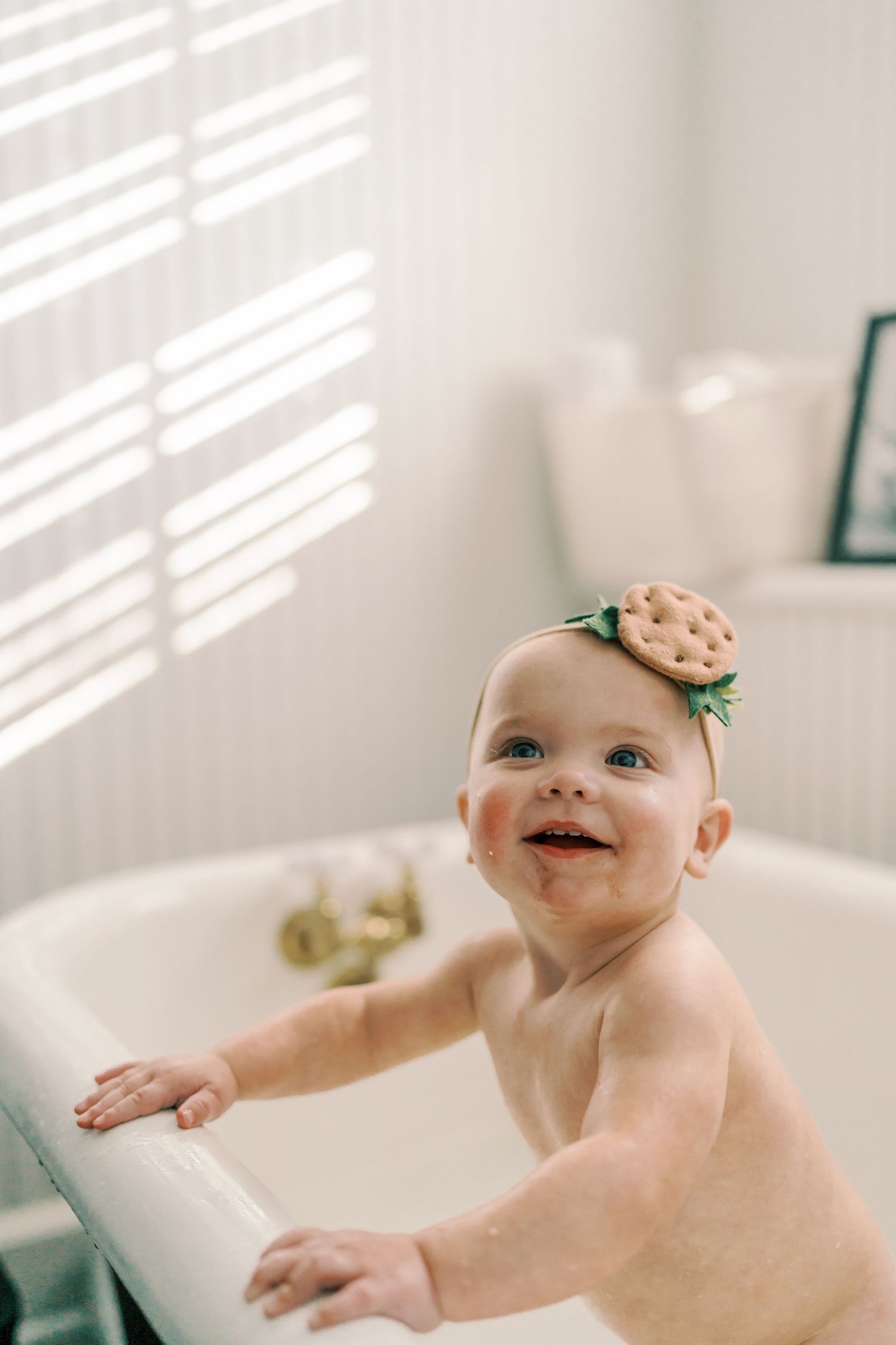 Baby splashes happily in bright white bathroom with sunlit window shadows.
