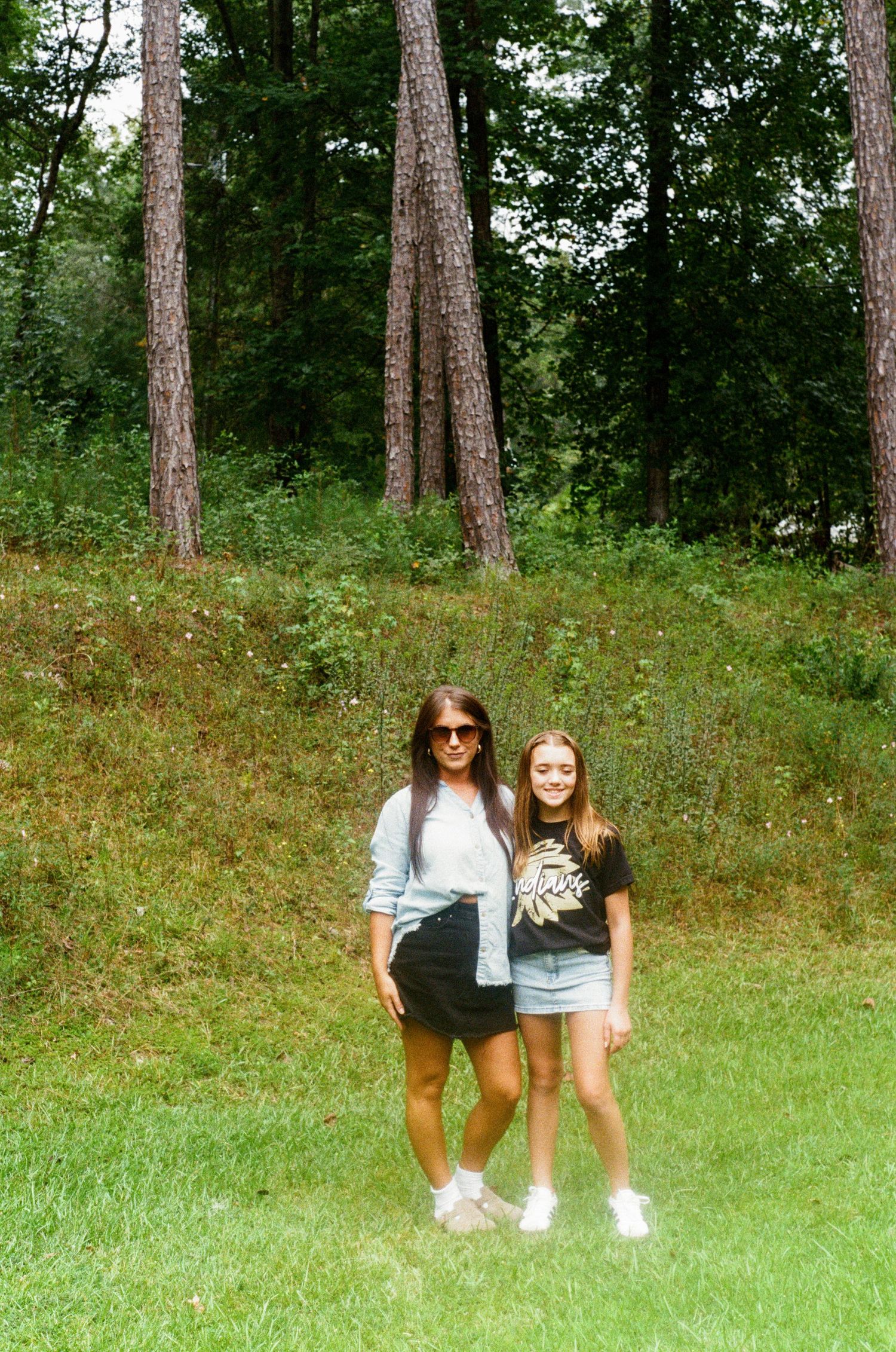 Two friends posing together in a wooded area with tall pine trees and grass in the background.