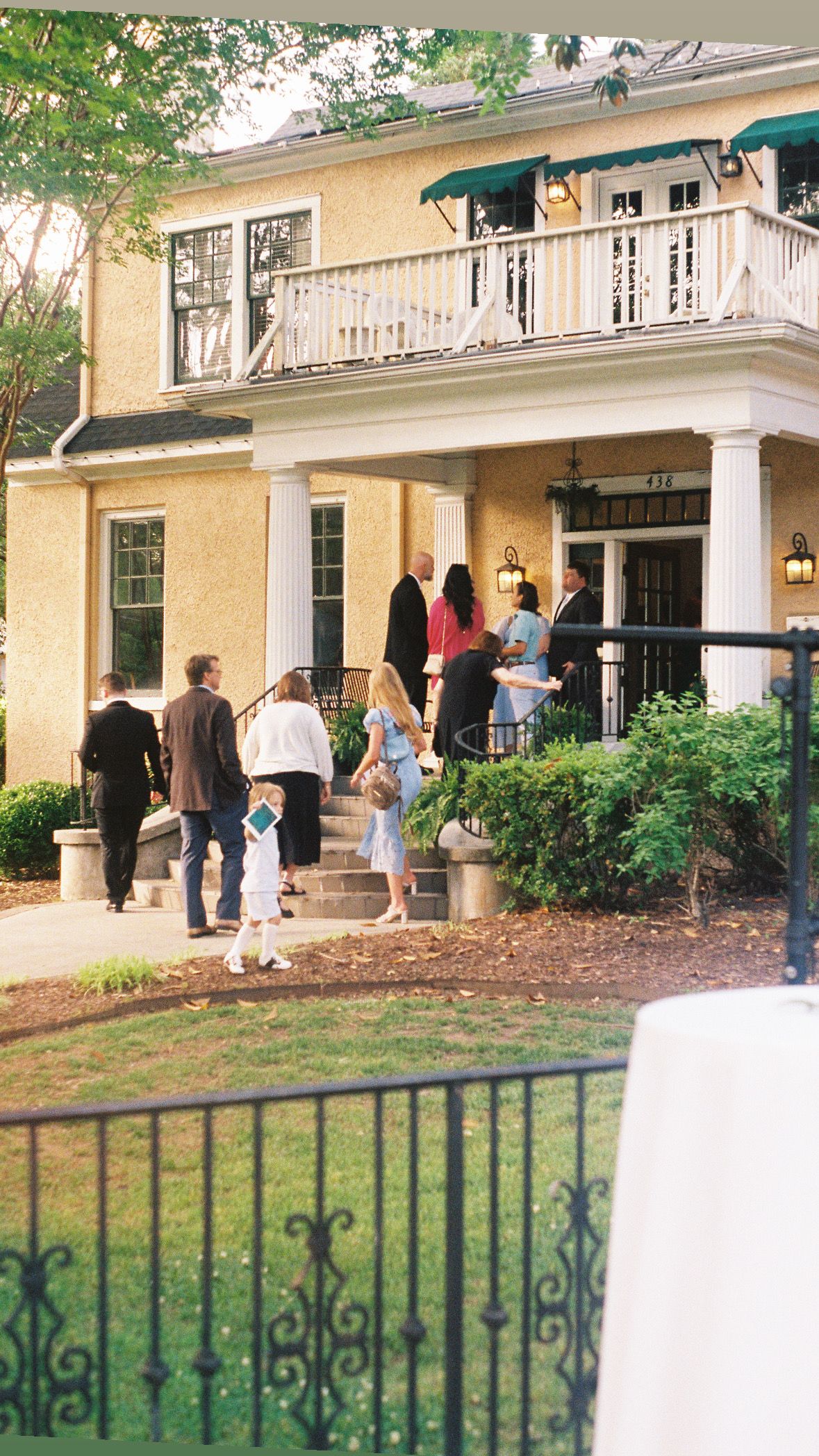 People gathering outside a large yellow colonial-style building with white columns.