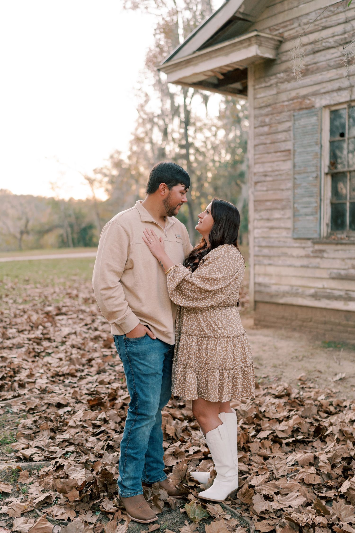 A couple in coordinating outfits poses lovingly near a rustic building surrounded by leaves.