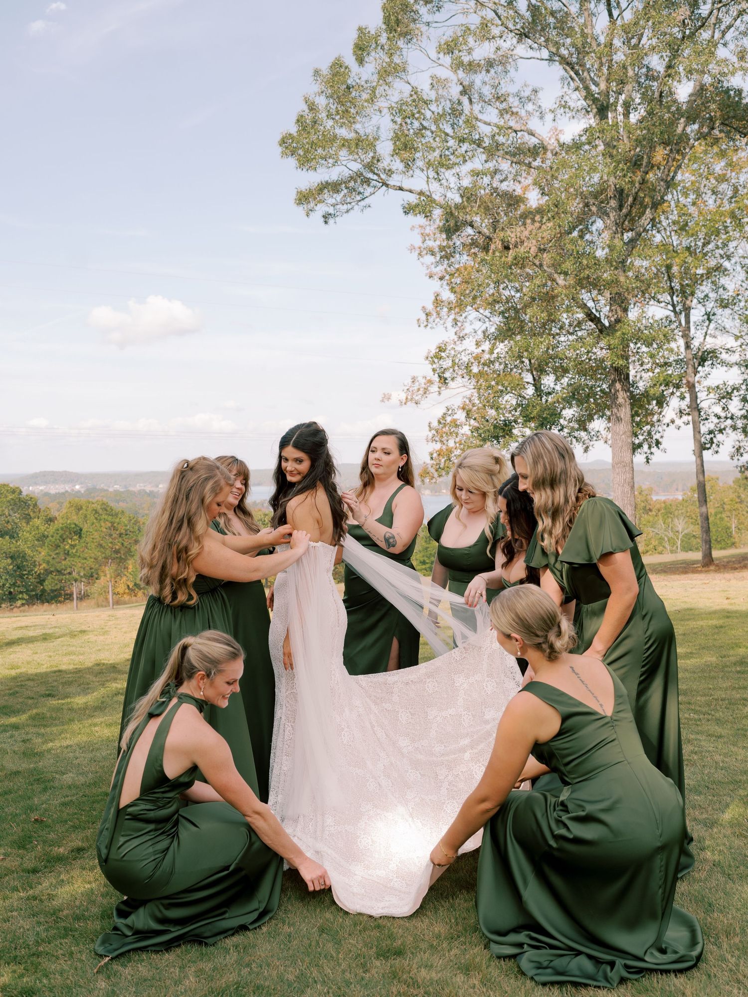 Bridesmaids in emerald green dresses helping with the wedding dress train outdoors.